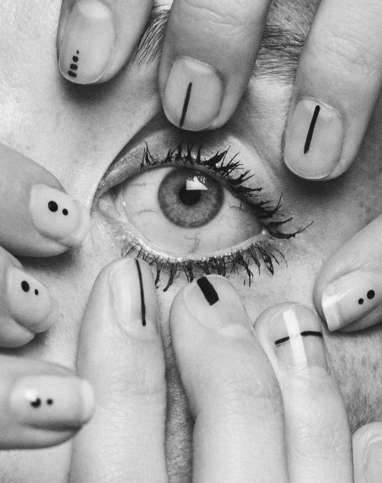 Black and white photo of a person's eye surrounded by fingers with various nail art designs, including dots, lines, and solid colors.