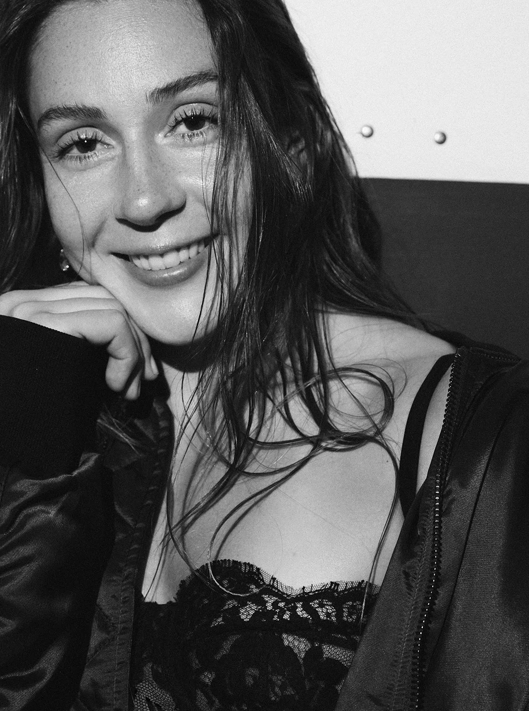 A smiling woman with wet hair, wearing a lace top and a jacket, resting her chin on her hand, in black and white.