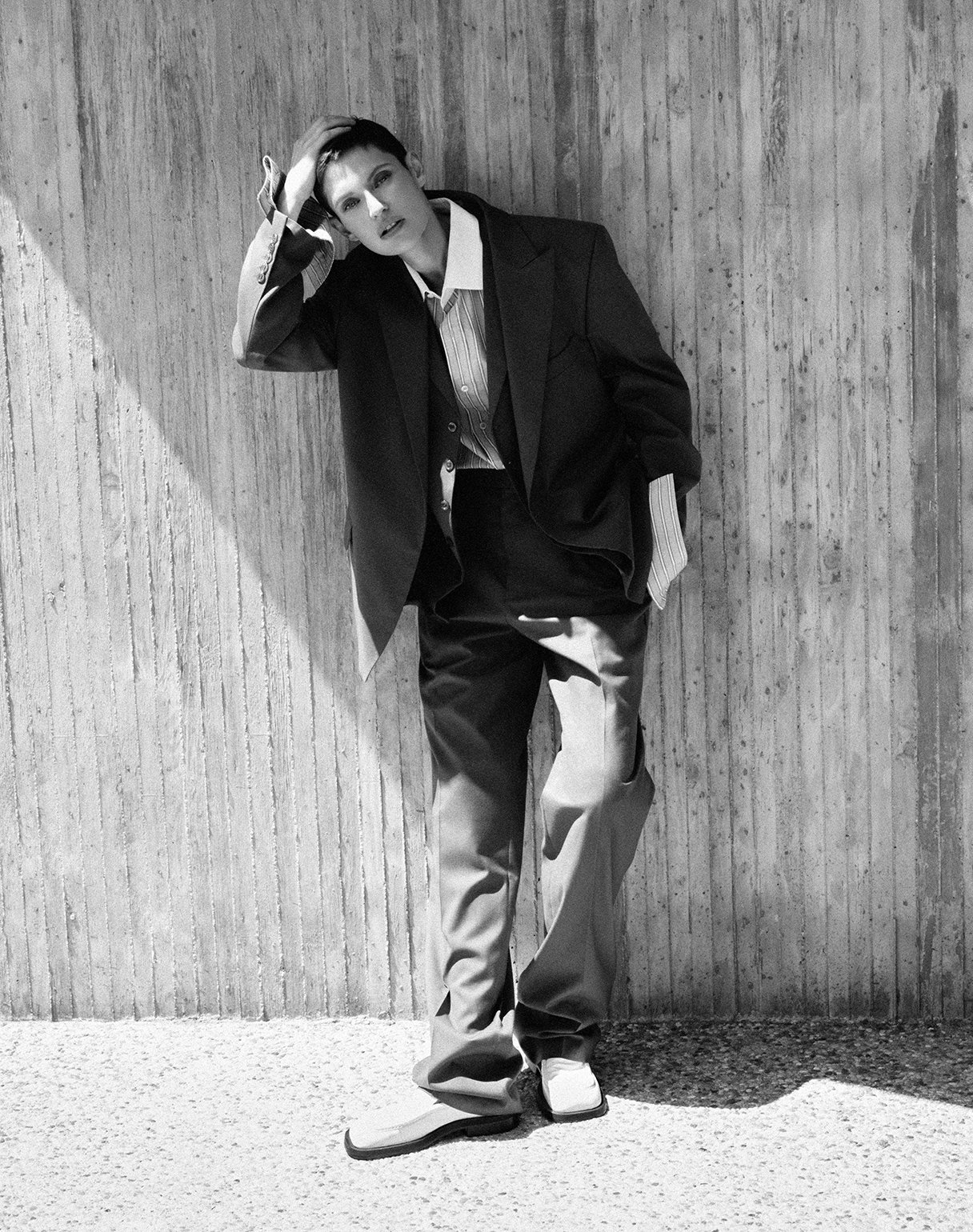 Black and white photo of a person with short hair, wearing a loose suit with a shirt, standing against a textured wall. They are leaning against the wall, with one hand on their head and the other in their pocket.