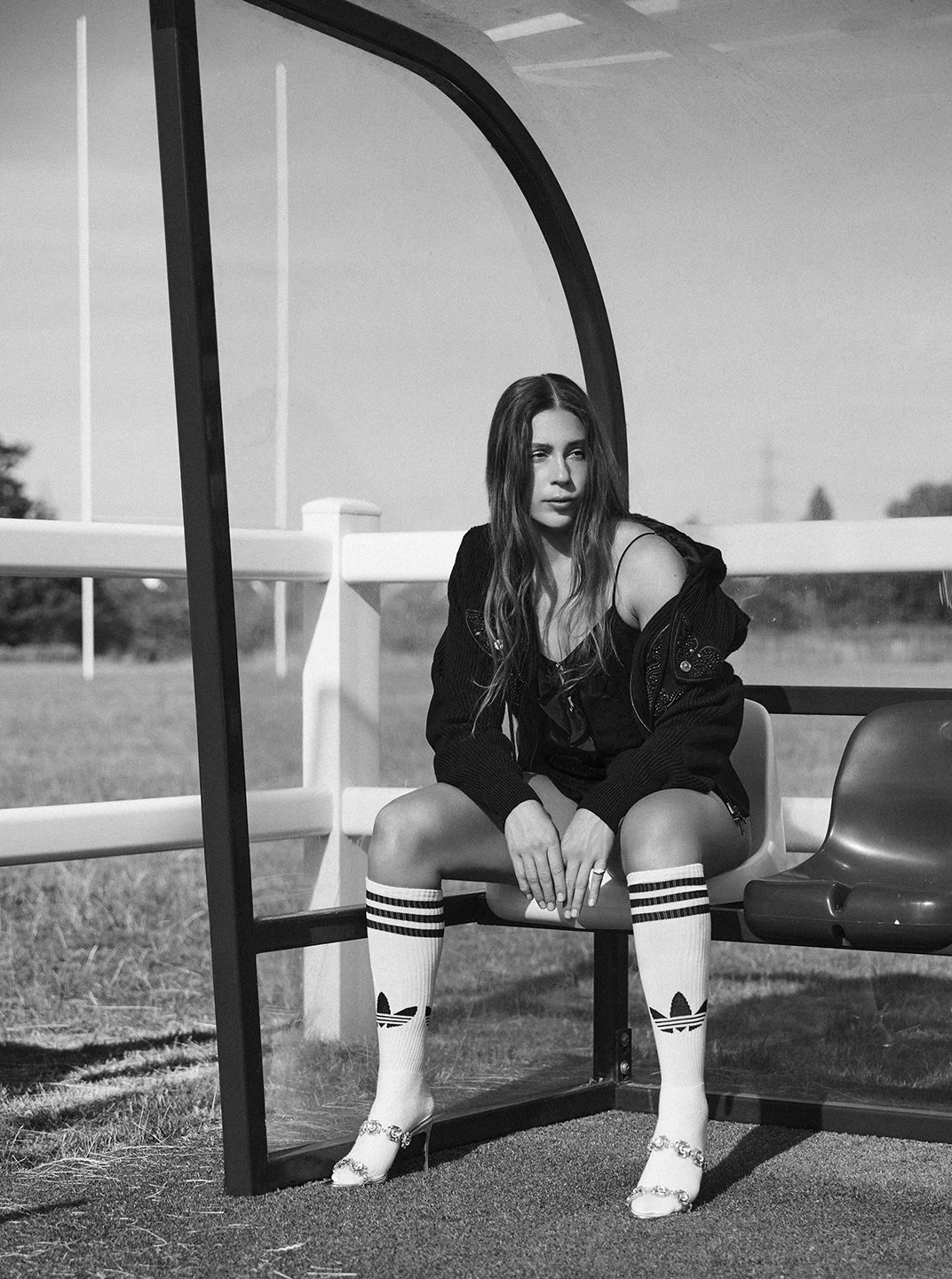 A woman sitting on a golf cart, wearing knee-high Adidas socks and high heels, in an outdoor setting with trees and a fence in the background.
