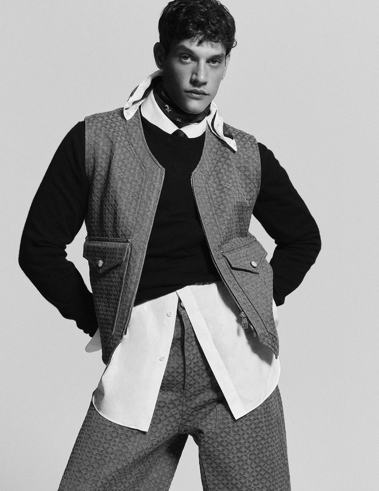 Black and white photo of a young man posing confidently, wearing layered clothing including a patterned vest over a sweater, with a shirt underneath, and patterned pants.