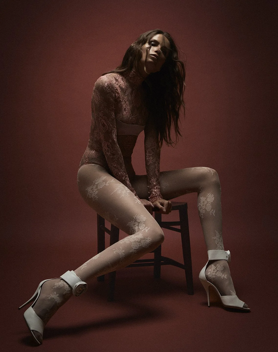 A woman in lace clothing and high heels sitting on a stool against a dark red background.