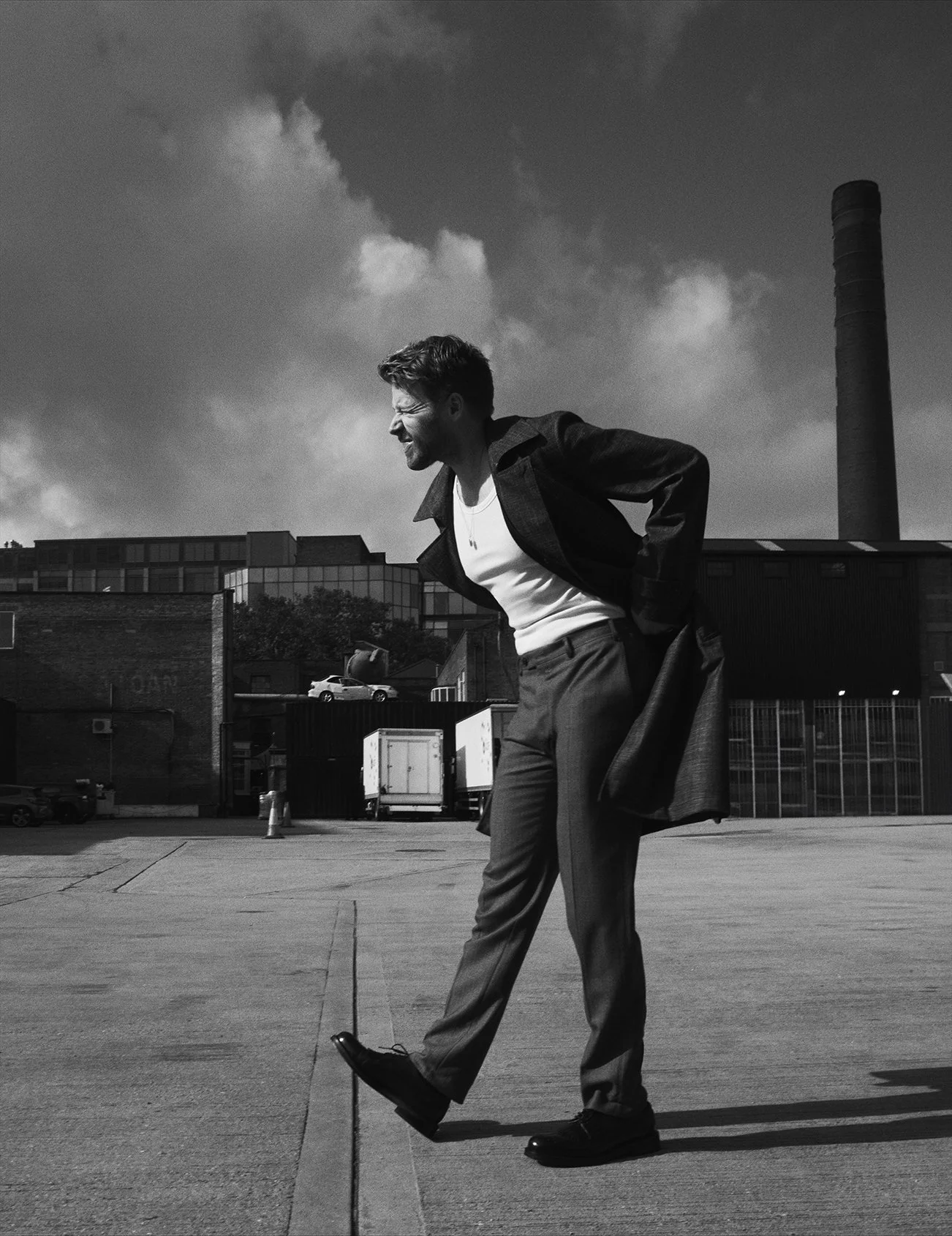 A young man in business attire at a parking lot, holding his lower back with a pained expression, with an industrial building and smokestack in the background, in black and white.
