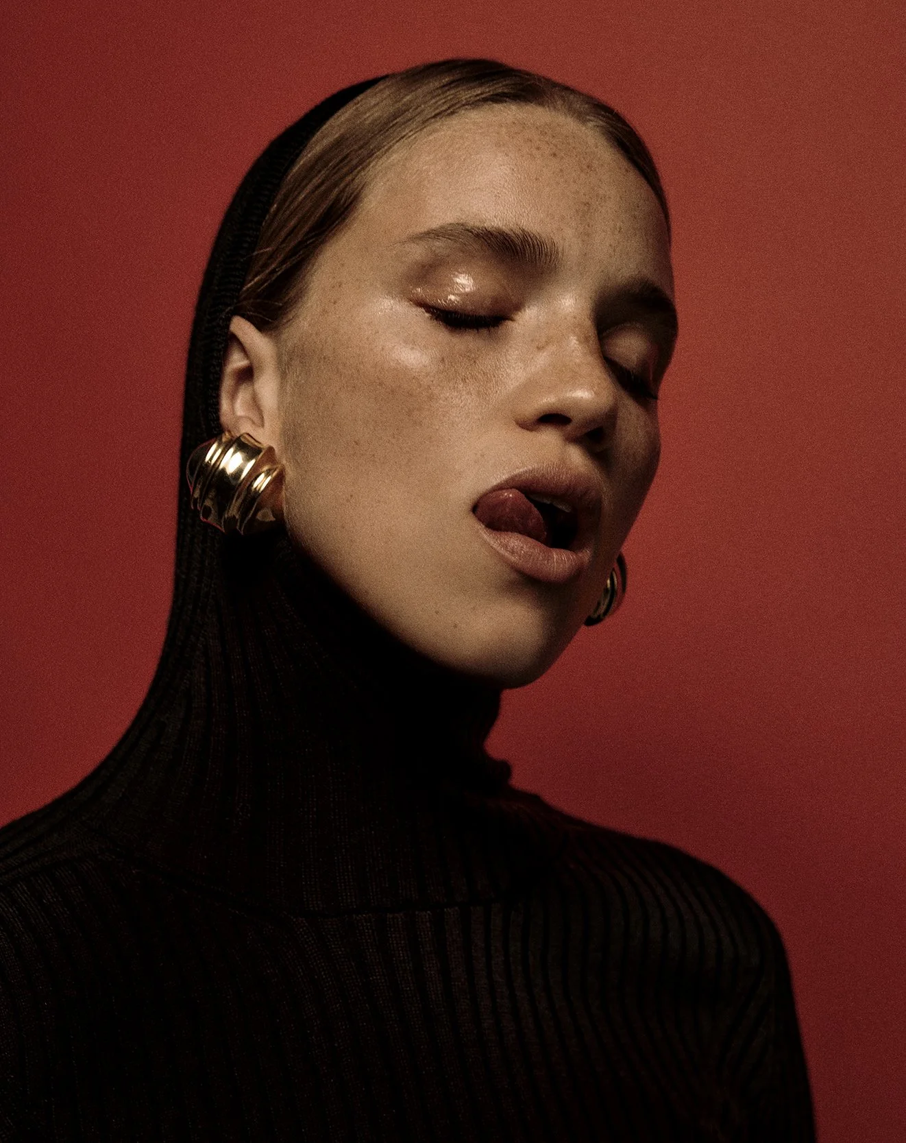 Close-up of a woman with freckles, wearing a black turtleneck, gold hoop earrings, with her eyes closed and tongue slightly out, against a red background.