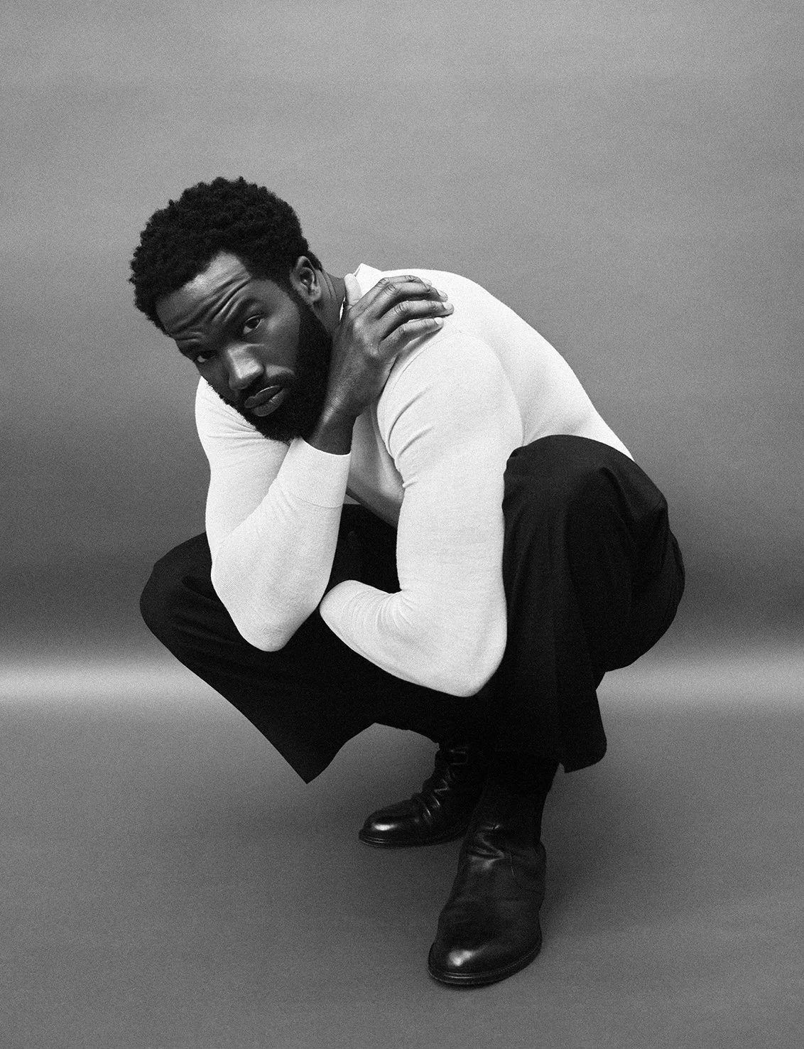 A Black man squatting in a studio, wearing a white long-sleeve shirt, black pants, and black boots, with his left hand on his shoulder and looking at the camera.