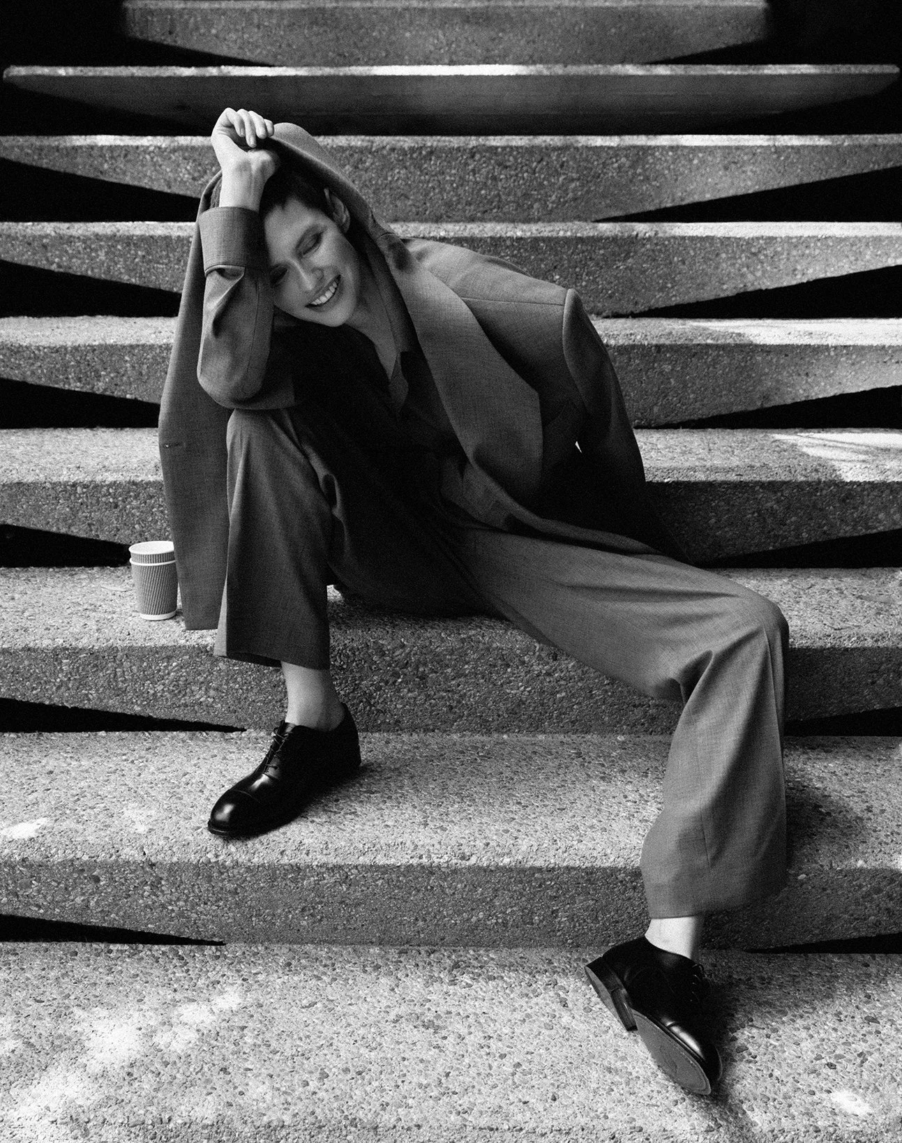A woman with short hair, dressed in a suit, sits on concrete steps, smiling and leaning her head on her hand, with a coffee cup beside her.