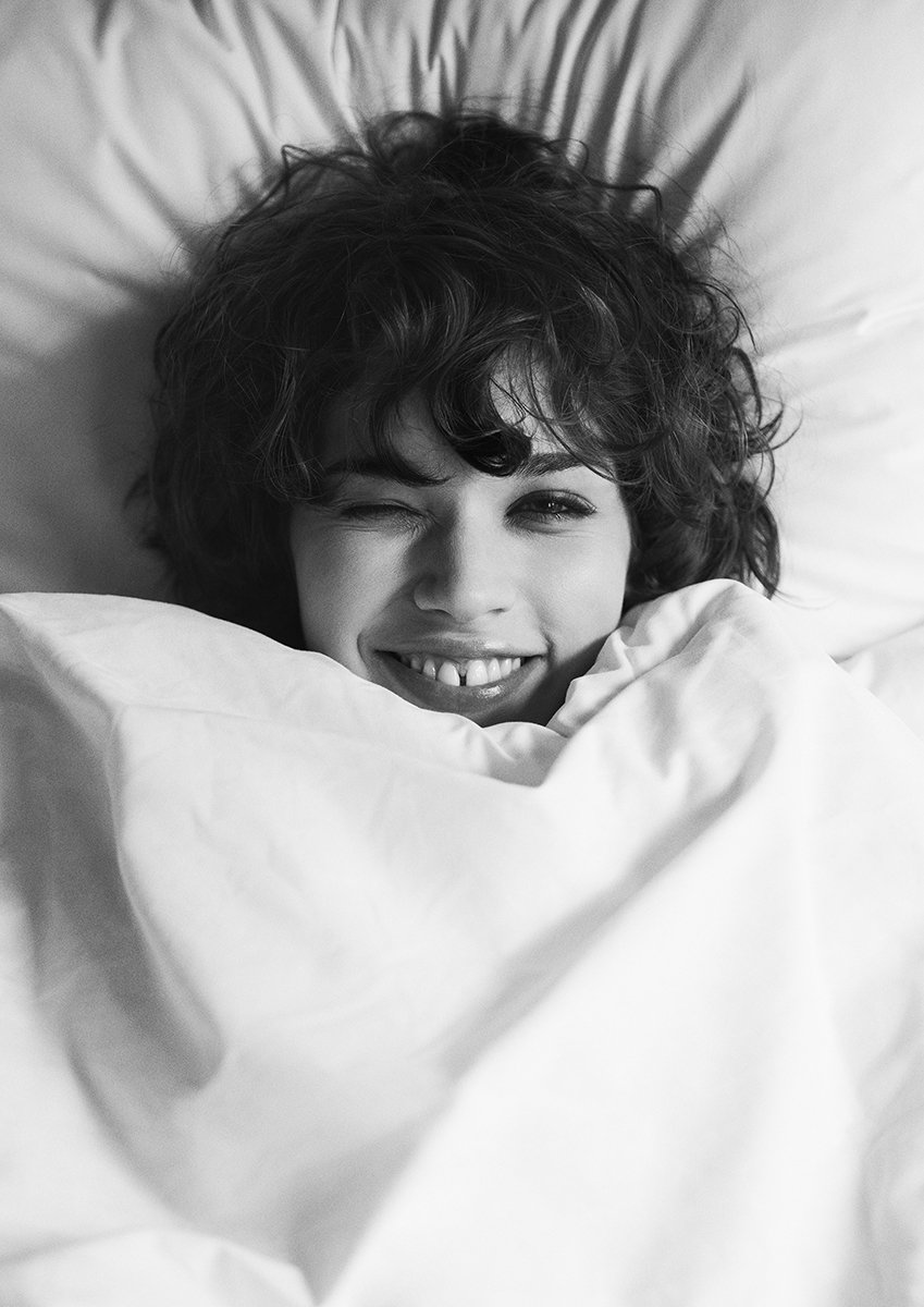 A black and white photo of a young child with curly hair, smiling and winking while lying in bed, partially covered with a blanket.