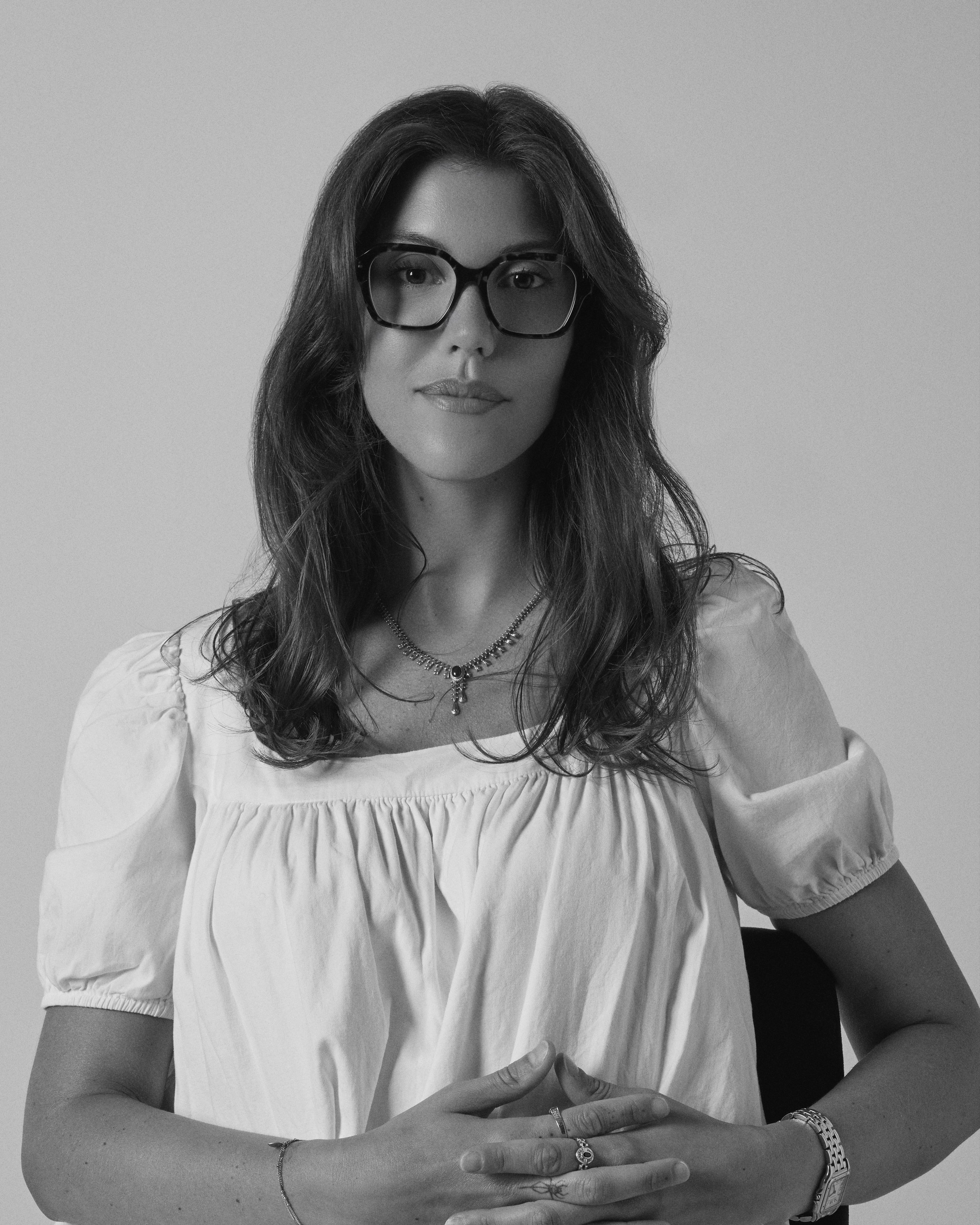 A woman with long wavy hair, wearing glasses, a white blouse, and jewelry, sitting against a plain background.