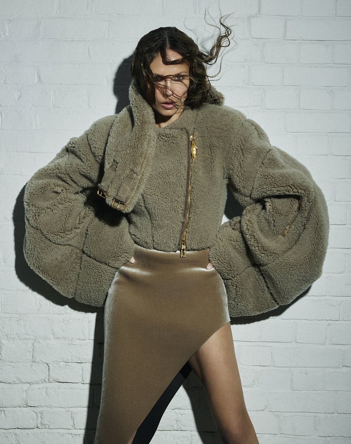 A woman with wavy brown hair, wearing a beige plush jacket and a metallic gold asymmetrical skirt, standing against a white brick wall with her hand on her head.