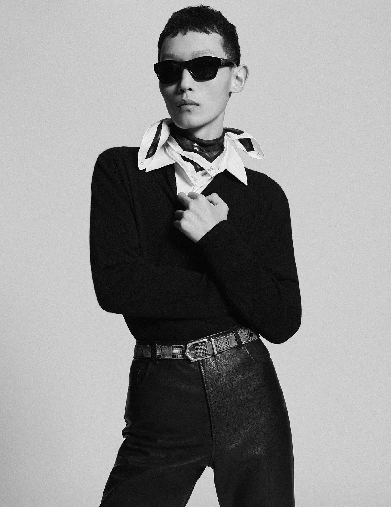 A black and white photograph of a young woman with short hair wearing sunglasses, a patterned scarf, a white collared shirt, a black sweater, and leather pants, posing with her hand on her chest.
