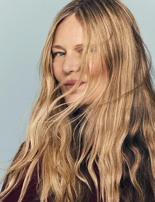 A woman with long, wavy blonde hair covering part of her face, winking and looking at the camera against a light blue background.