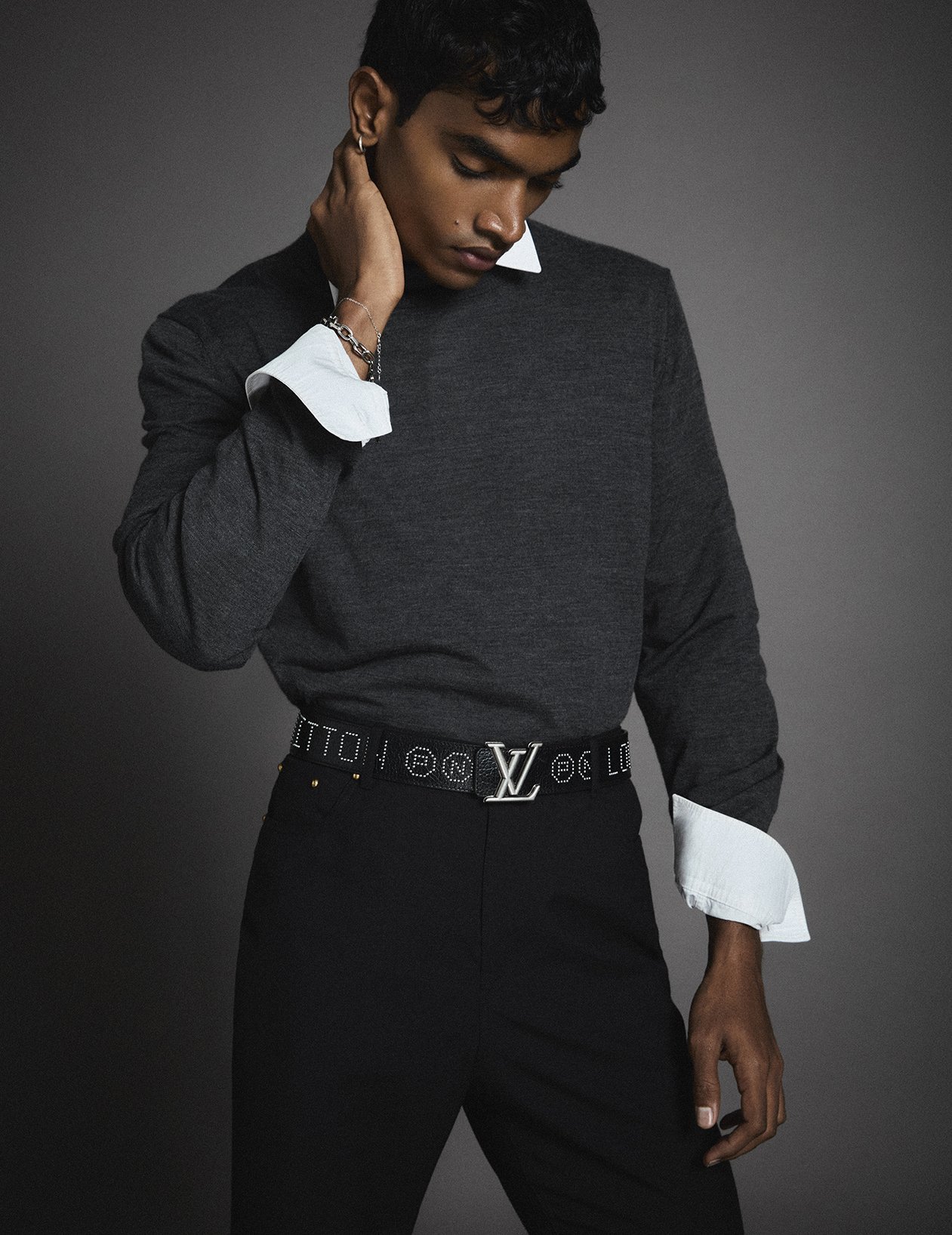 A young man wearing a dark gray sweater with a white shirt underneath, black pants, and a Louis Vuitton belt, posing against a dark background.