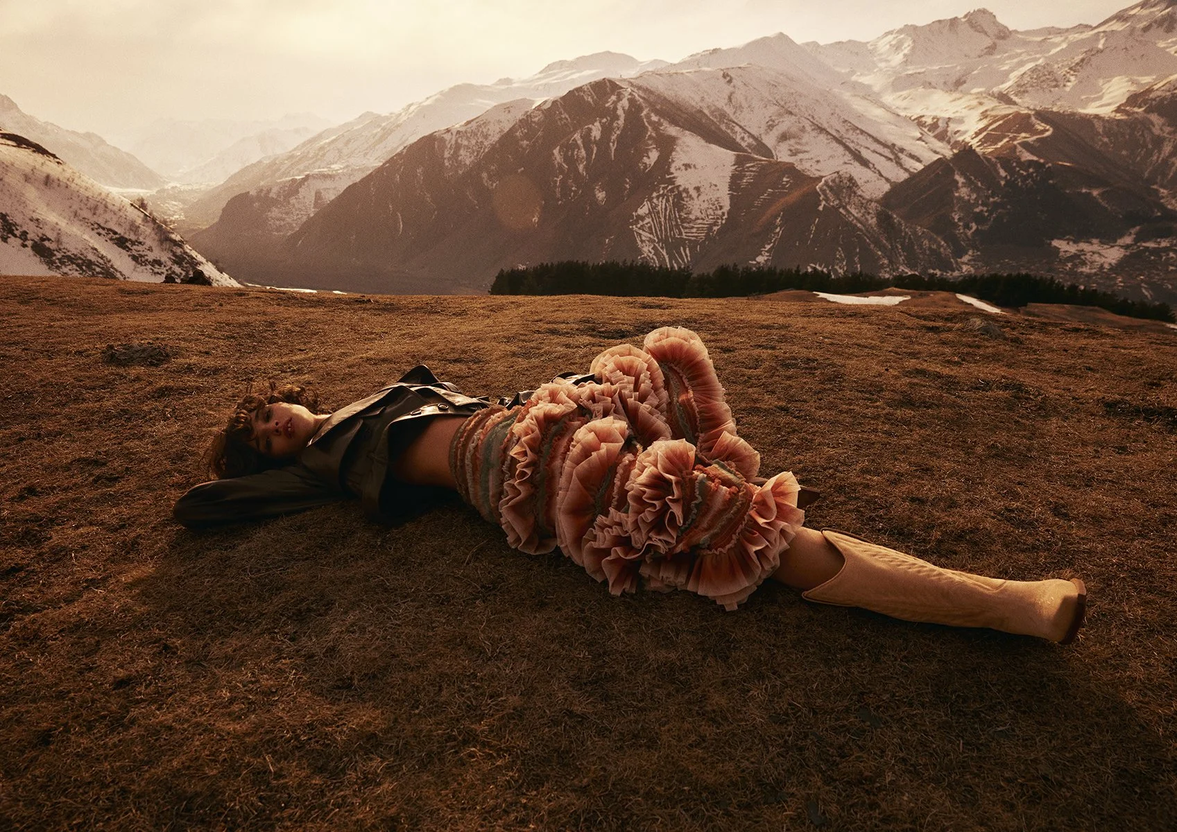 A woman lying on the ground in a mountainous landscape, with snow-capped mountains in the background and a cloudy sky, wearing a brown jacket, a ruffled multicolored skirt, and beige boots.