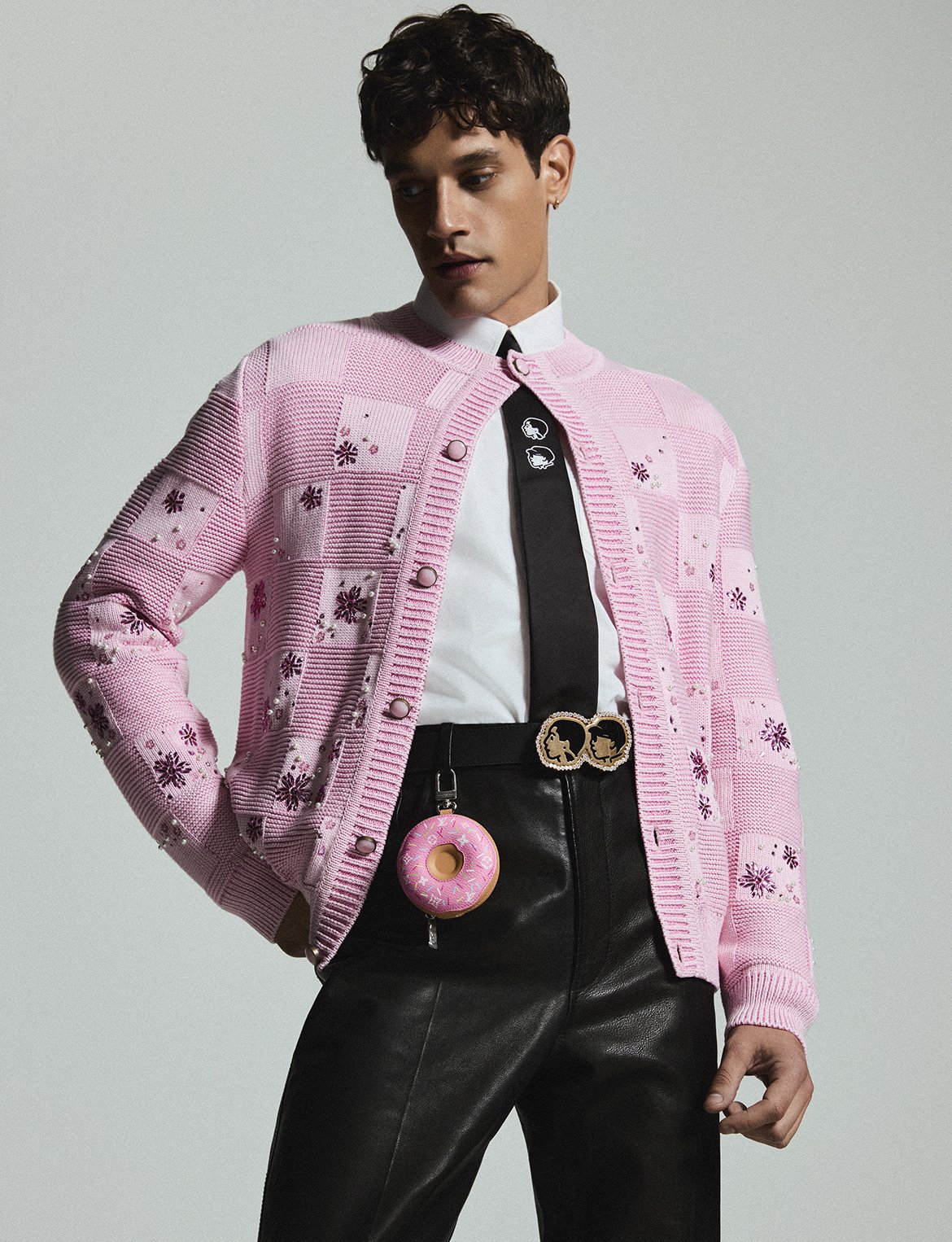 A person wearing a pink patterned cardigan over a white shirt and black tie, with black leather pants and a decorative belt featuring two faces. They are accessorized with a donut-shaped keychain hanging from their pocket and small earrings, standing