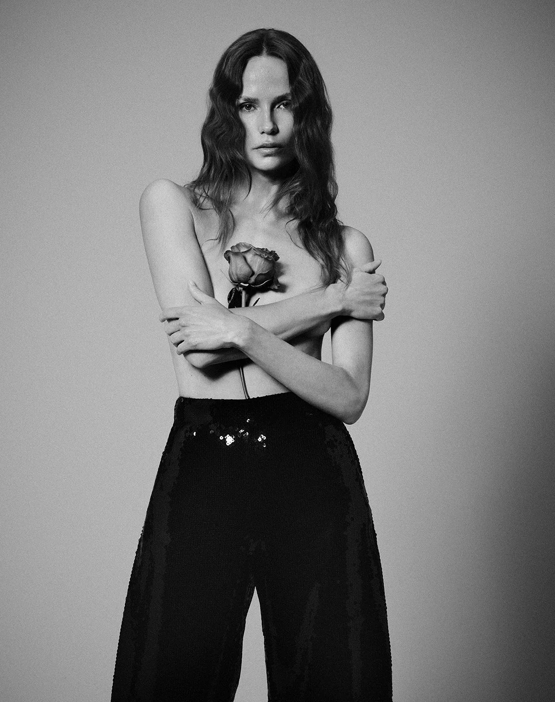 Black and white photo of a woman with wavy hair, covering her chest with her arms, holding a rose close to her chest, wearing high-waisted pants, and looking at the camera.