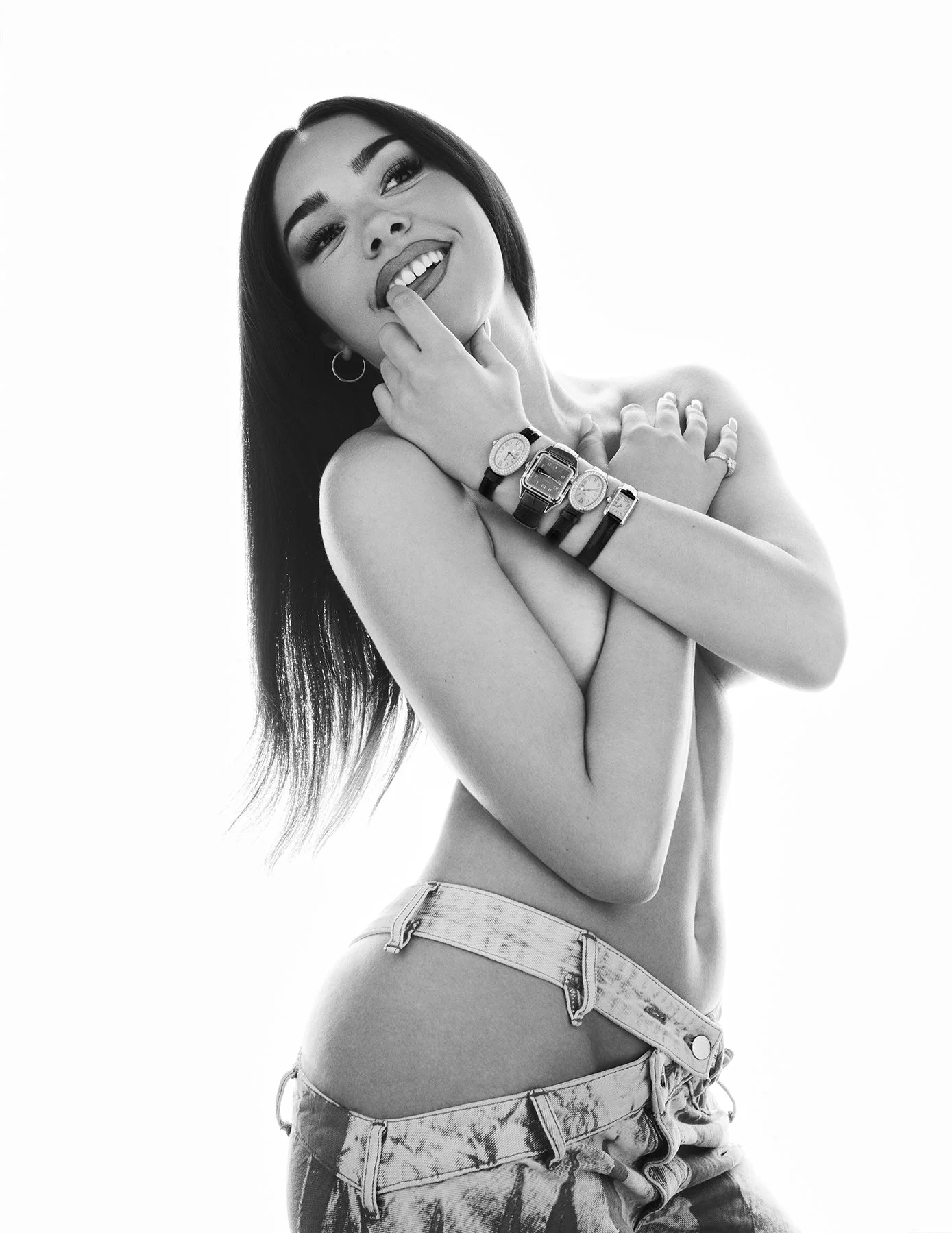 Black and white photo of a smiling woman with long dark hair, wearing multiple watches, with her hand touching her lips, and wearing unbuttoned jeans.