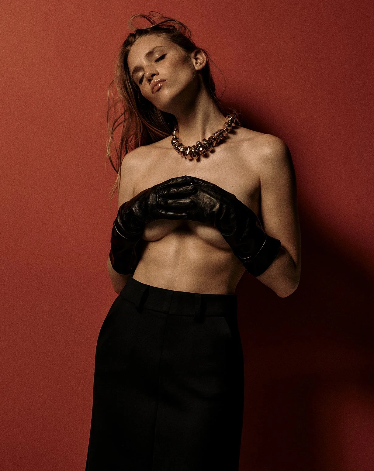 A woman poses topless against a red background, covering her chest with her arms and wearing black gloves, a statement necklace, and black pants.