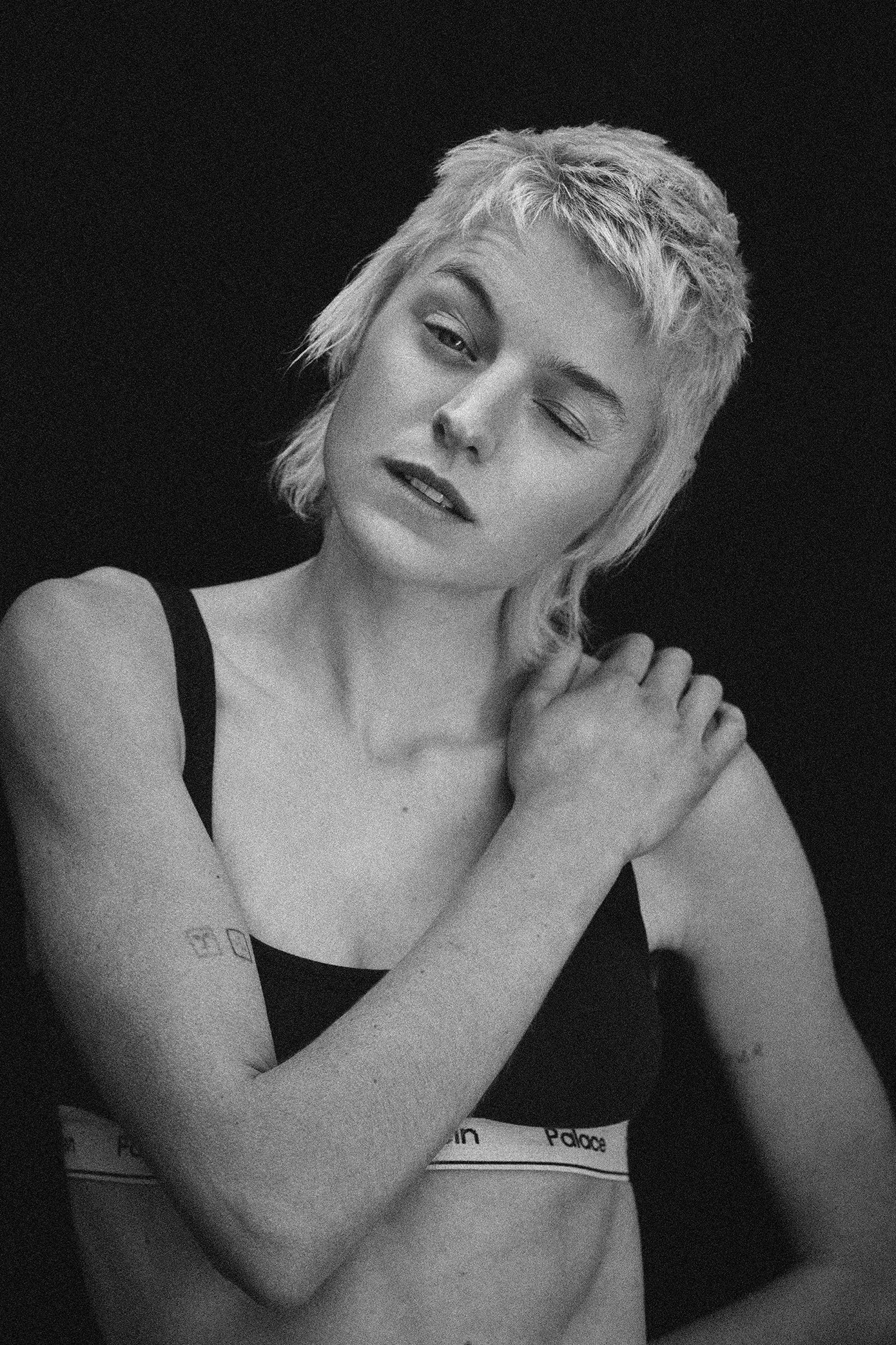 Black and white portrait of a woman with short, tousled blonde hair, one eye closed, wearing a sports bra, with her right hand resting on her shoulder.