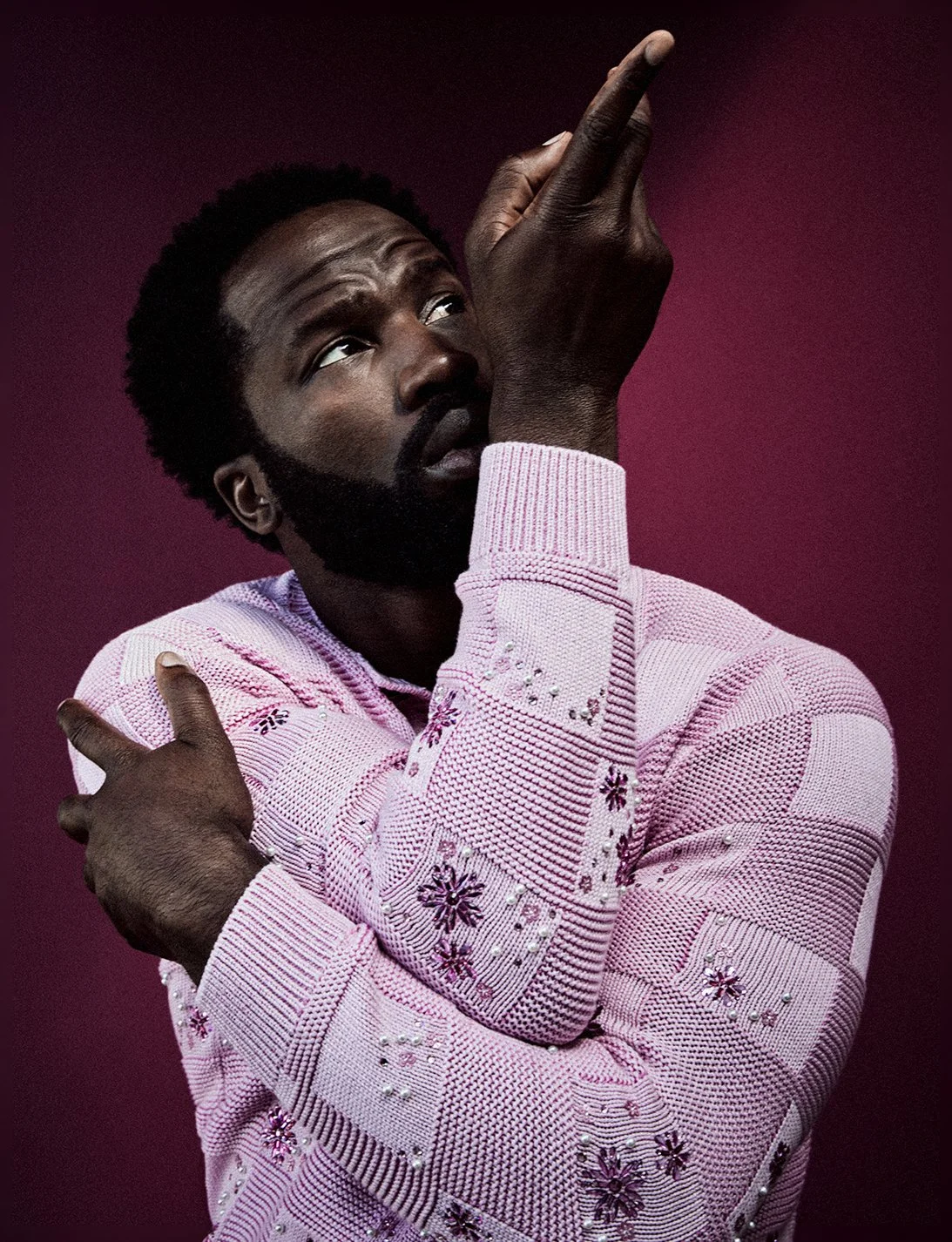 A man with a beard and curly hair, wearing a pink knit sweater with floral embroidery and pearl details, poses against a dark pink background. He is pointing upward with his right hand and crossing his left arm over his chest.