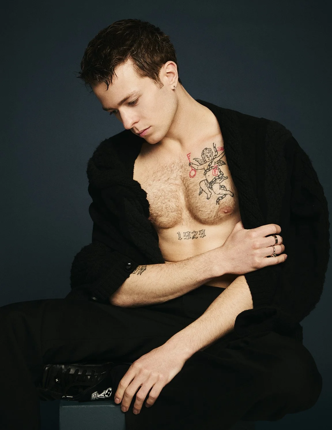 A young man with brown hair looking down, partially unbuttoned black shirt, revealing tattoos on chest and abdomen, seated against a dark background.