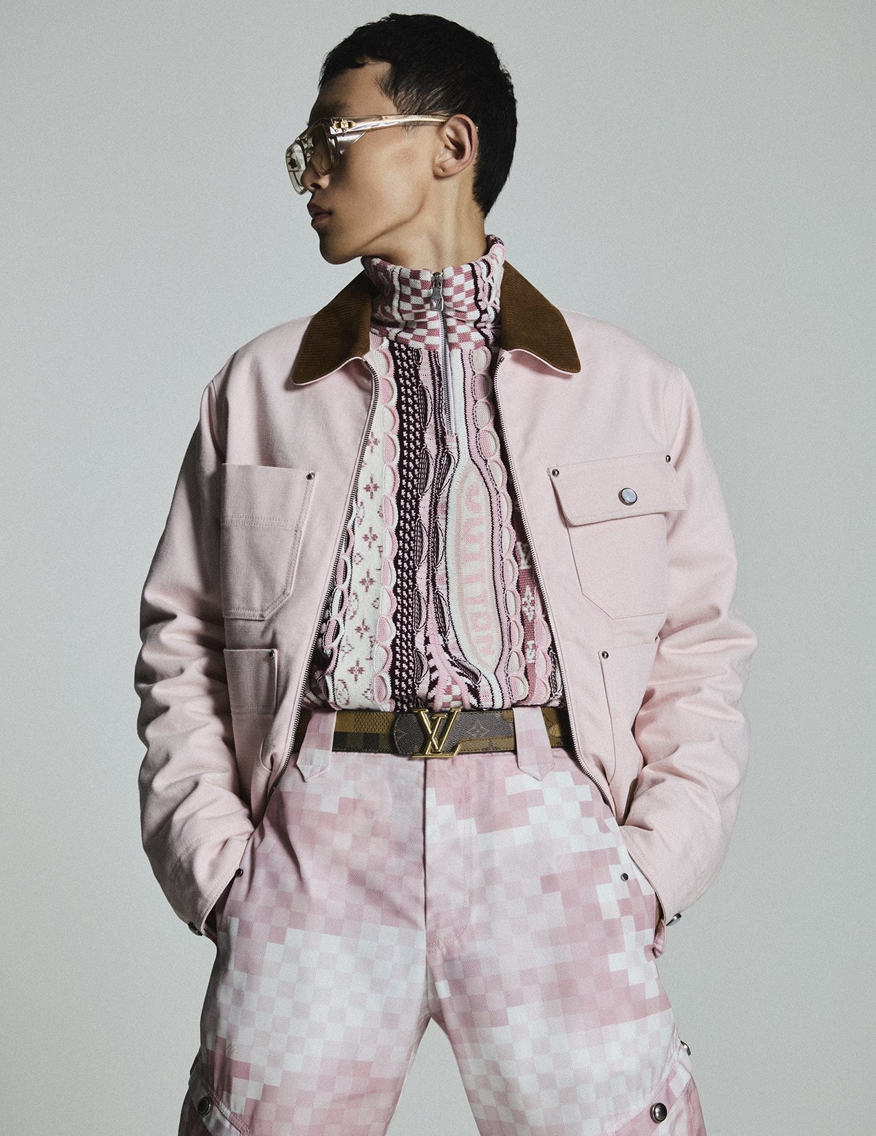 A person wearing a pink jacket, patterned shirt, and pink checkered pants with hands in pockets, looking sideways, wearing glasses.