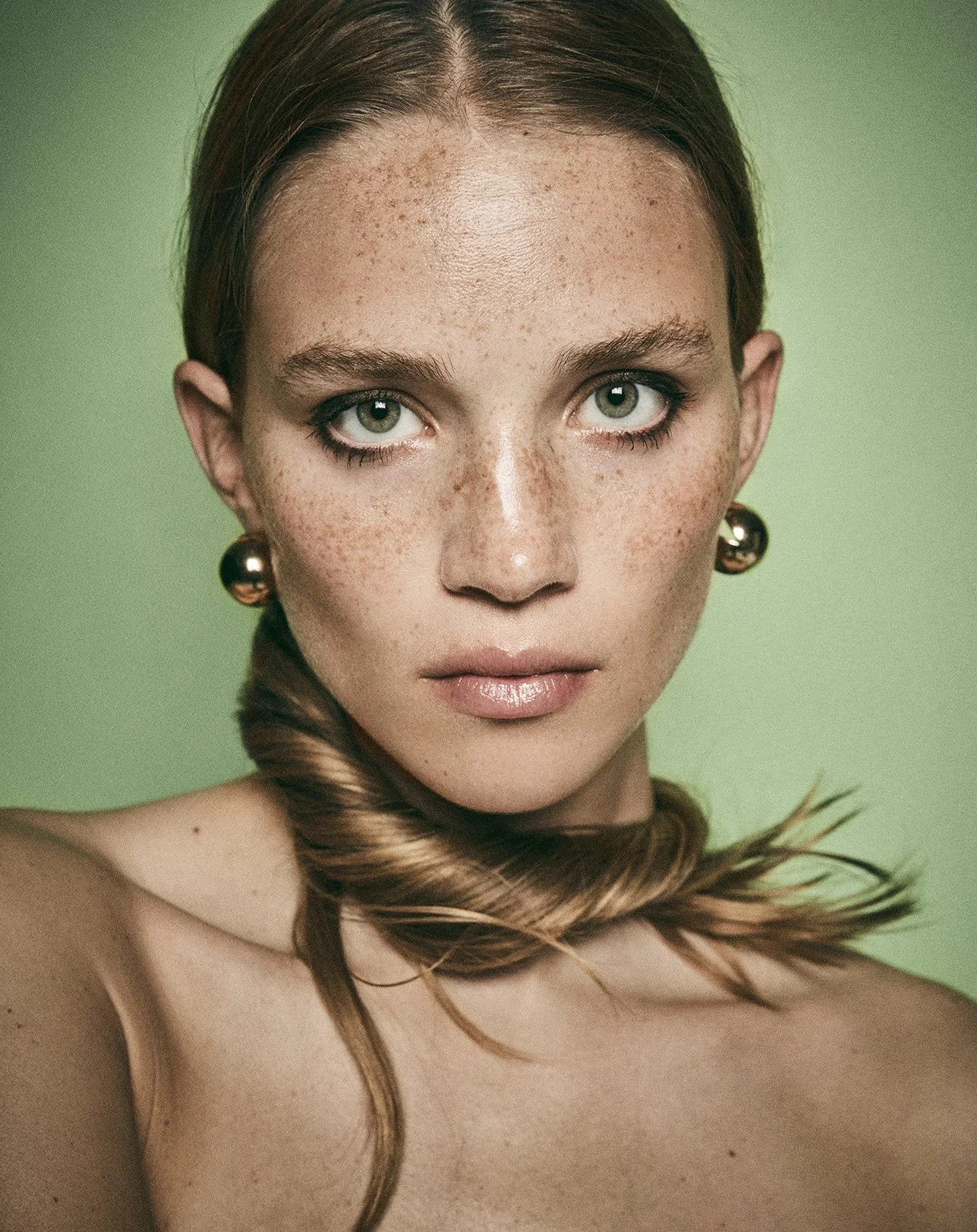 Close-up portrait of a woman with freckles, green eyes, brown hair, and gold earrings against a green background.