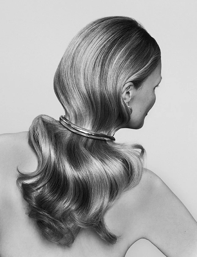 Black and white photo of a woman with long, wavy, shiny hair styled in waves, wearing a necklace and hoop earring, with her face turned to the right.
