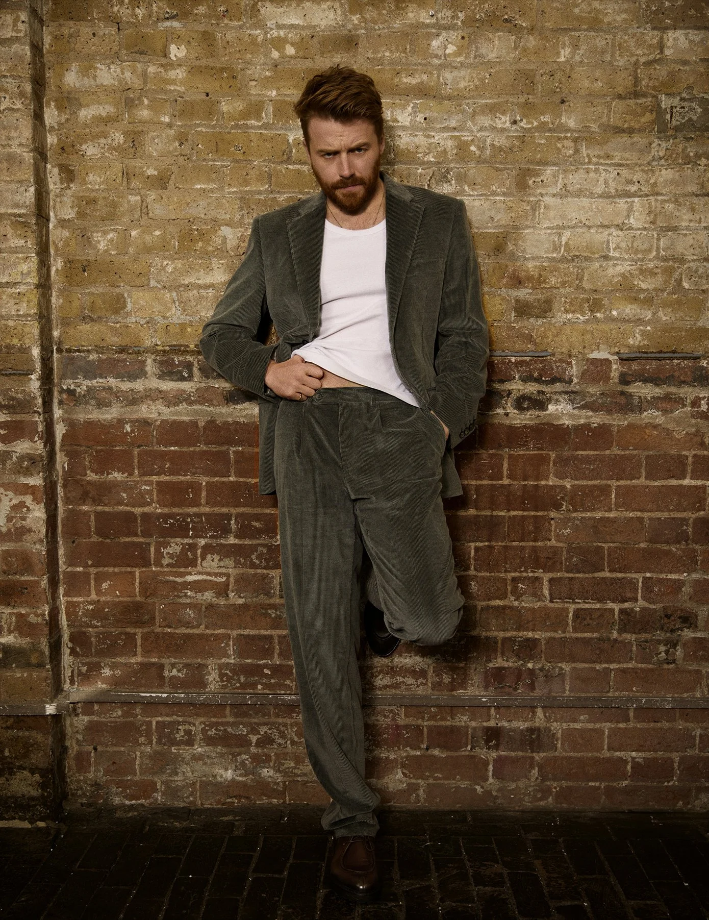 Man with red hair and beard leaning against a brick wall, wearing a gray suit and white t-shirt, with one hand in pocket and the other adjusting his waistband.
