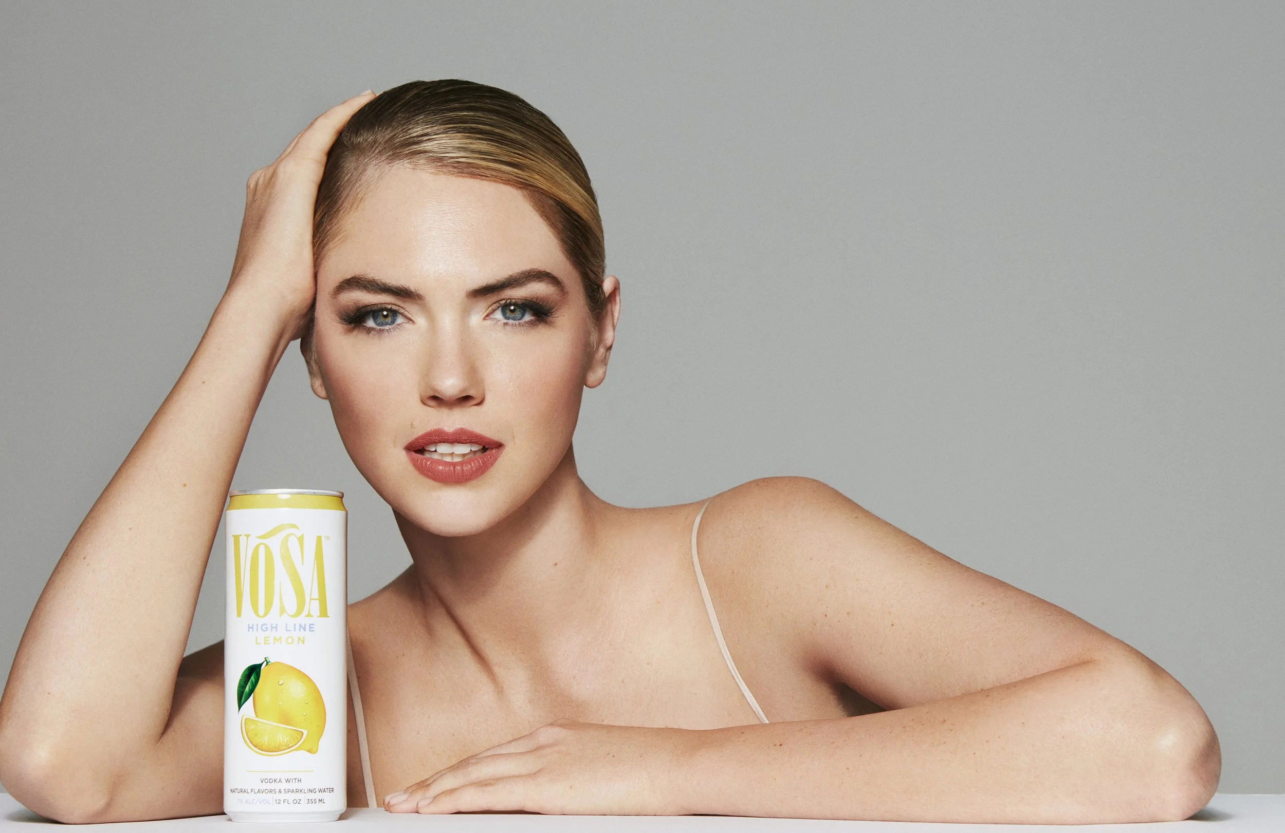 A woman with blonde hair, light makeup, and light skin gazes at the camera, resting her head on her left hand. She is next to a can of VOSA High Line Lemon flavored alcoholic beverage, with her right arm resting on a surface.