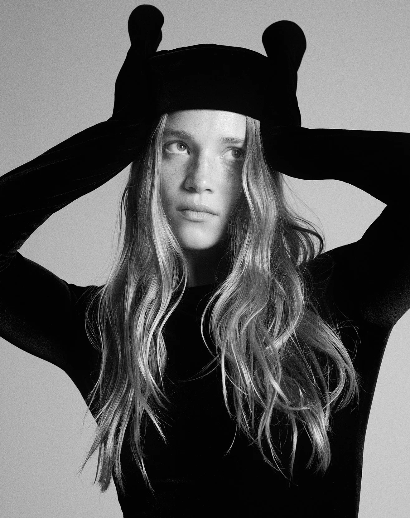 Black and white portrait of a woman with long wavy hair, wearing a dark top and a hat with bear ears, holding the hat with both hands, looking thoughtfully upward.