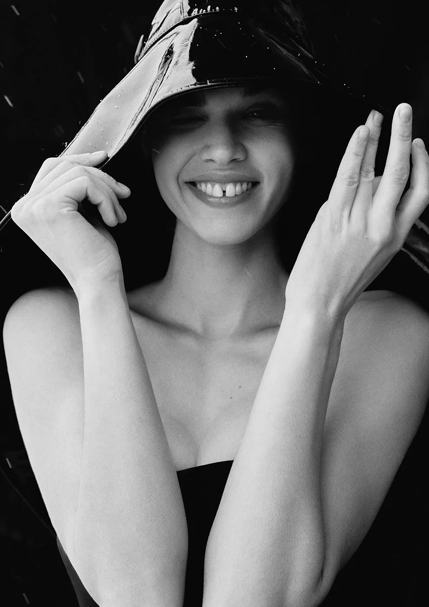 Black and white photo of a smiling woman wearing a raincoat with a hood, holding the hood with one hand and touching her face with the other, raindrops on her raincoat.