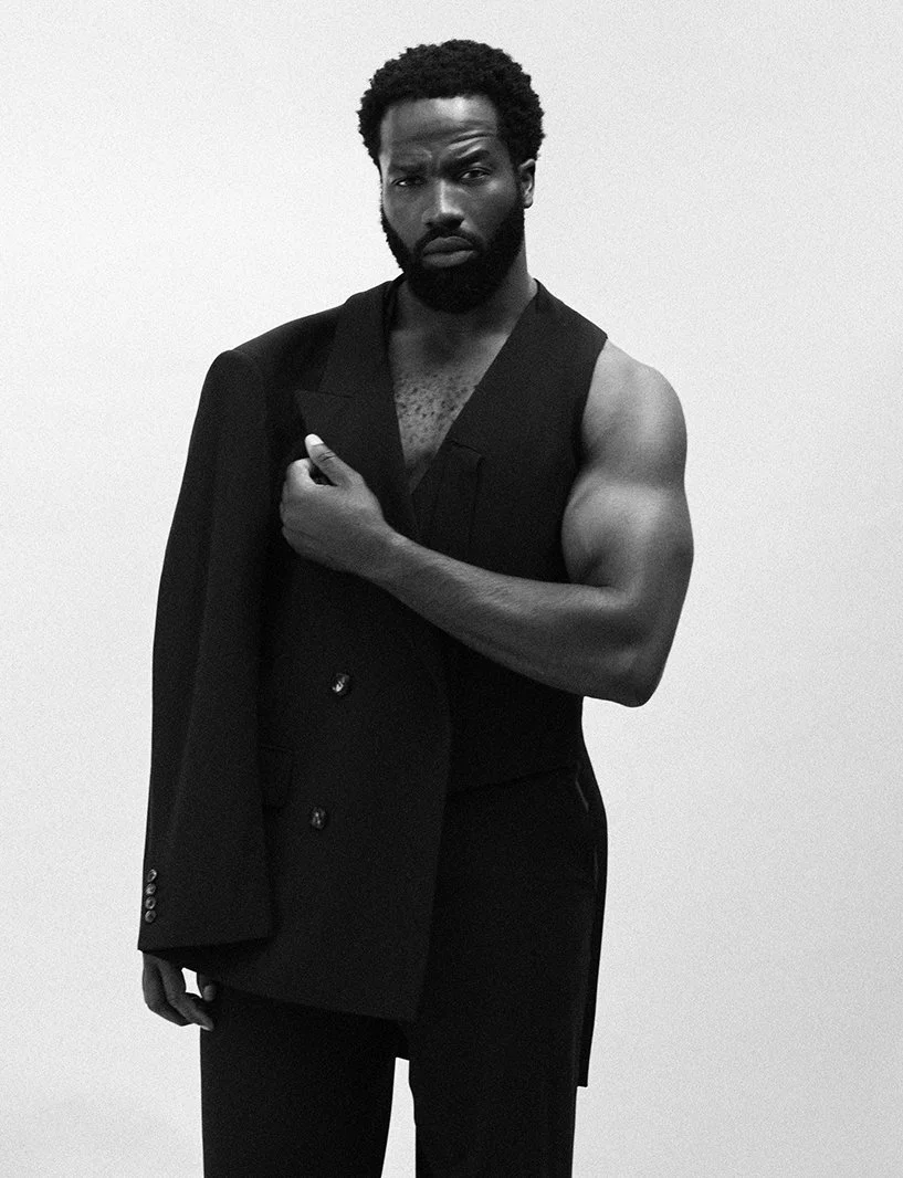 Black and white portrait of a man with a beard, standing against a plain white background. He is shirtless with a sleeveless vest, holding a blazer over his shoulder with one hand, and has a serious expression.