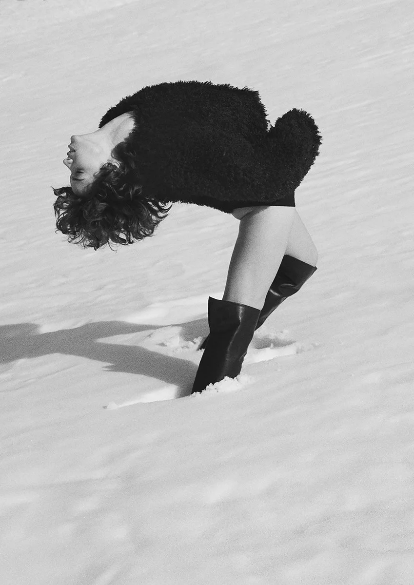 A woman with curly hair bent over in the snow, wearing a black coat and high boots, with her head upside down.