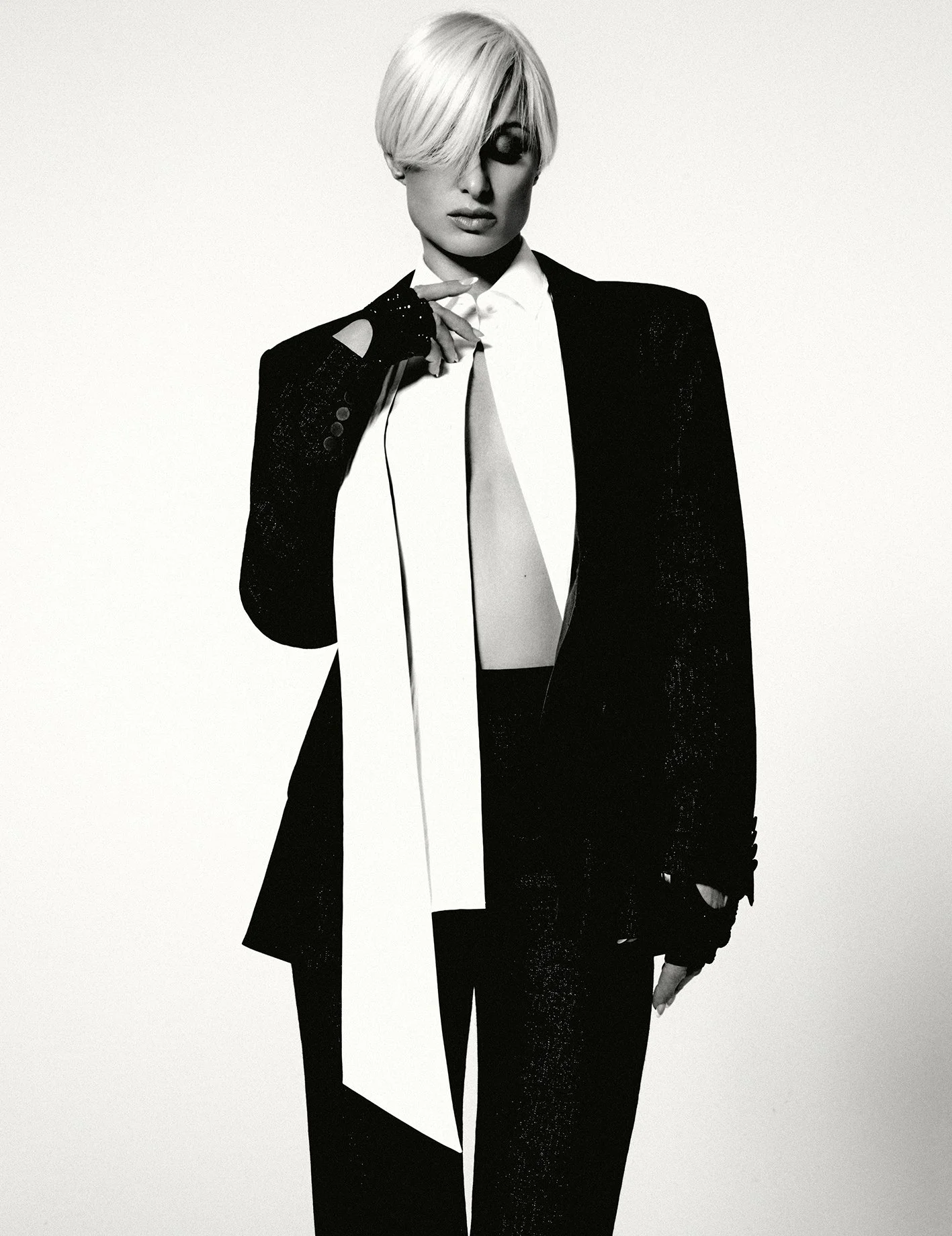 A woman with short blonde hair wearing a black suit with a white shirt, holding a microphone and standing against a plain background.