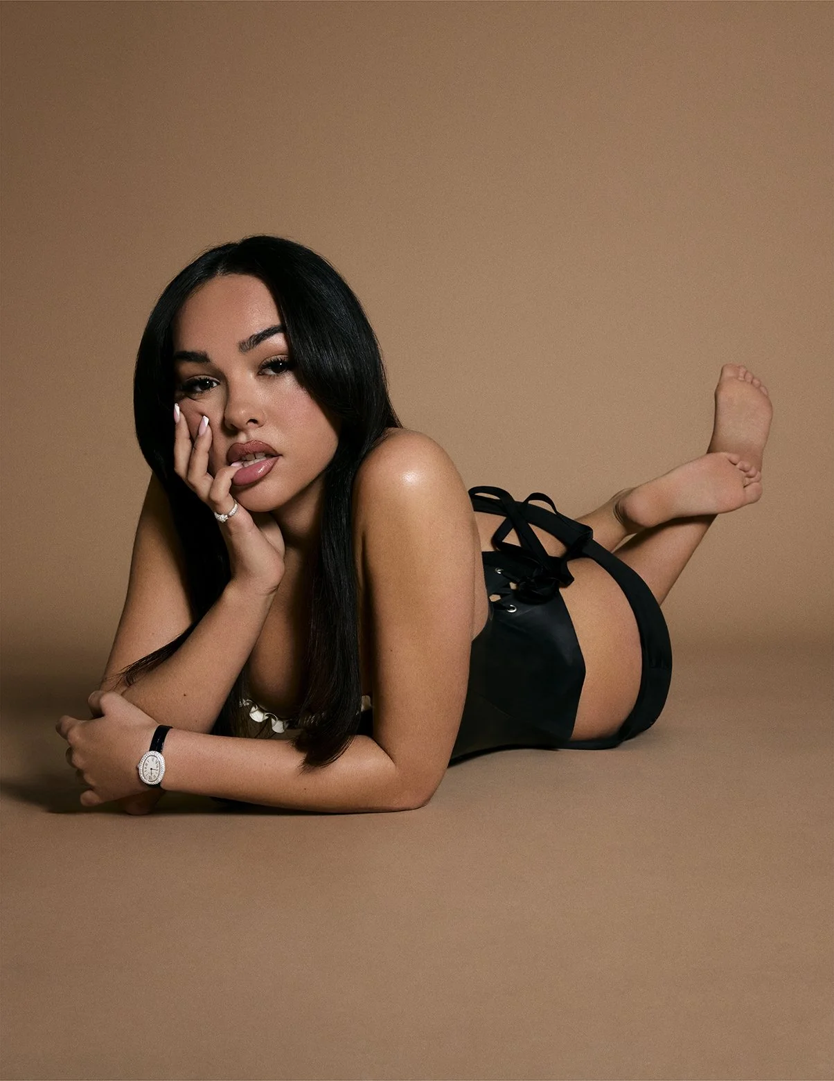 A woman with long black hair lying on her stomach on a tan background. She is wearing black lingerie, a watch, and a ring, and is looking at the camera with a relaxed expression.