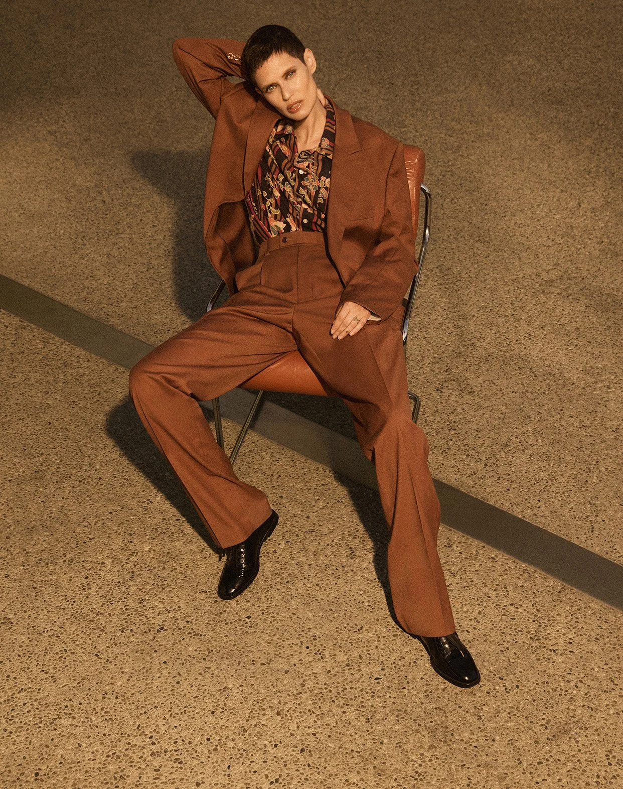 A woman with short dark hair wearing a brown suit and black shoes, sitting on a brown chair against a textured beige floor, with one arm resting on her head.