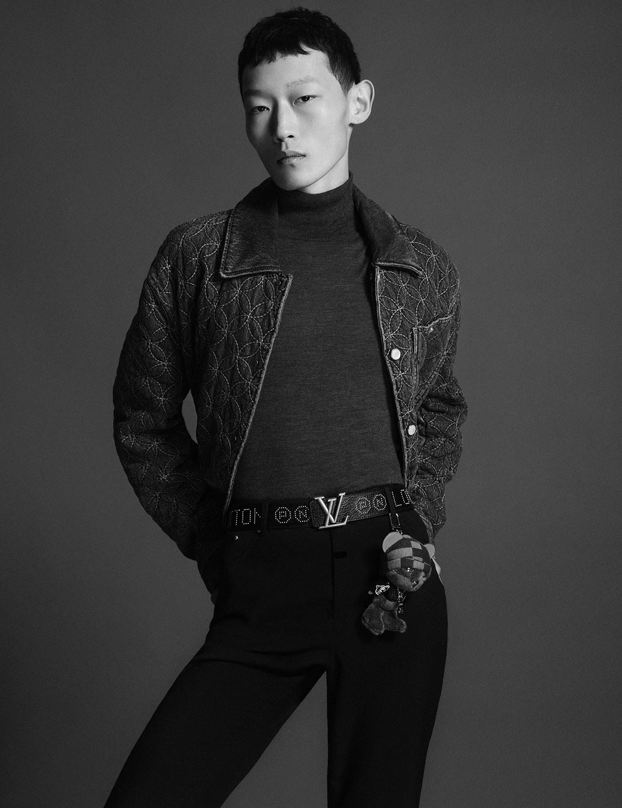 A black and white studio portrait of an Asian man with short dark hair, wearing a patterned leather jacket over a turtleneck, black trousers, and a Louis Vuitton belt.