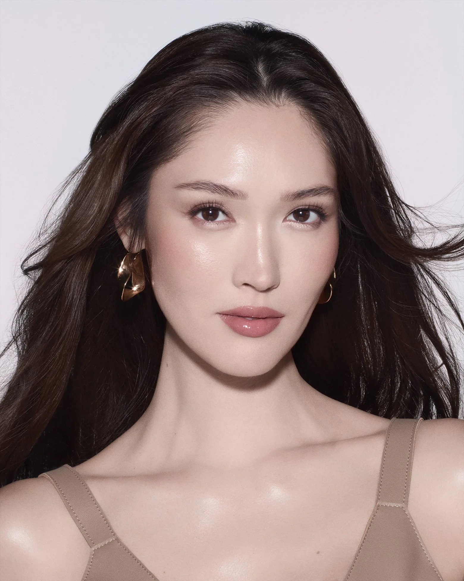 A woman with long, dark brown hair, wearing gold earrings and a beige top, looking directly at the camera against a plain white background.