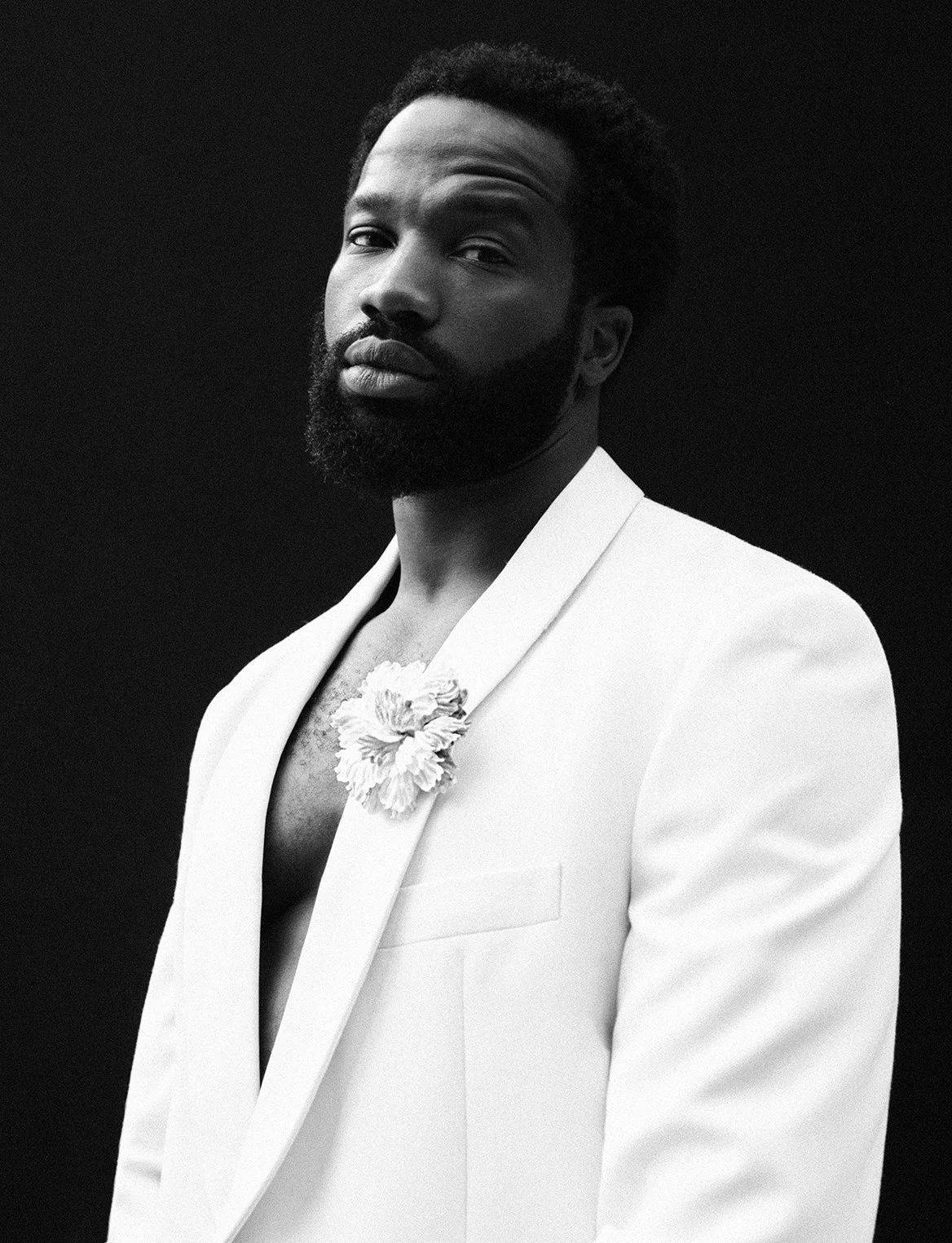 A black and white portrait of a man with a beard wearing a white suit jacket with a floral embellishment on the lapel, posing against a dark background.