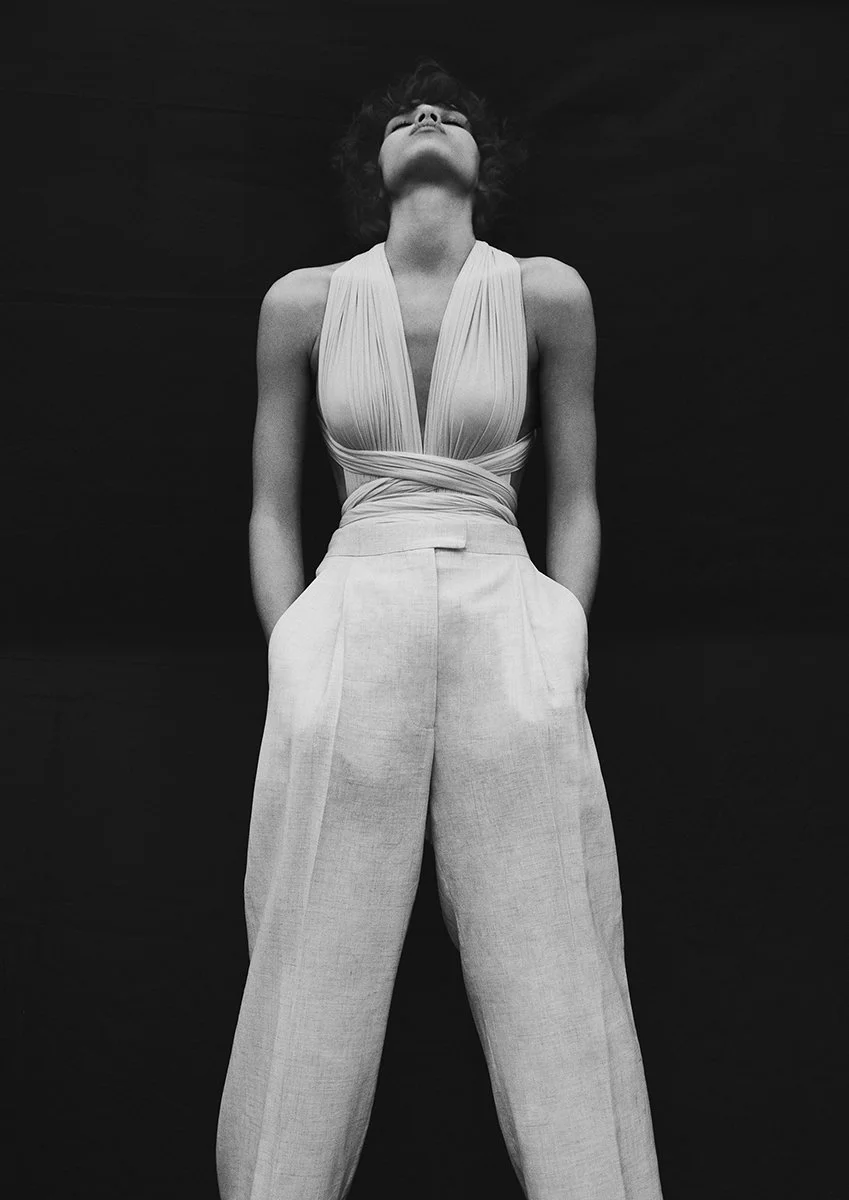 A black and white photo of a woman standing against a dark background, looking upwards with her hands behind her back. She is wearing a sleeveless top with a deep V-neck and high-waisted pants.