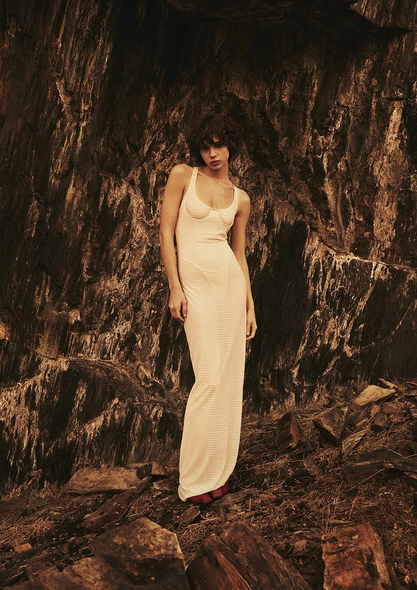 A woman in a long, light-colored dress standing against a rocky, rugged outdoor background.