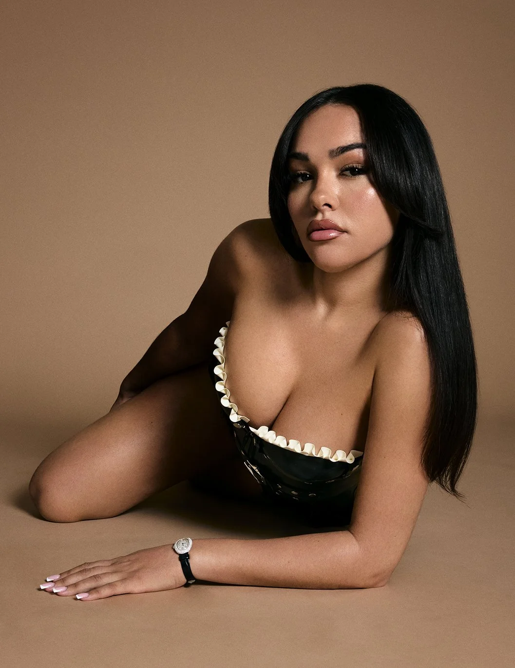 A woman with long black hair lying on her side, wearing a strapless black top with white ruffle trim, against a beige background.