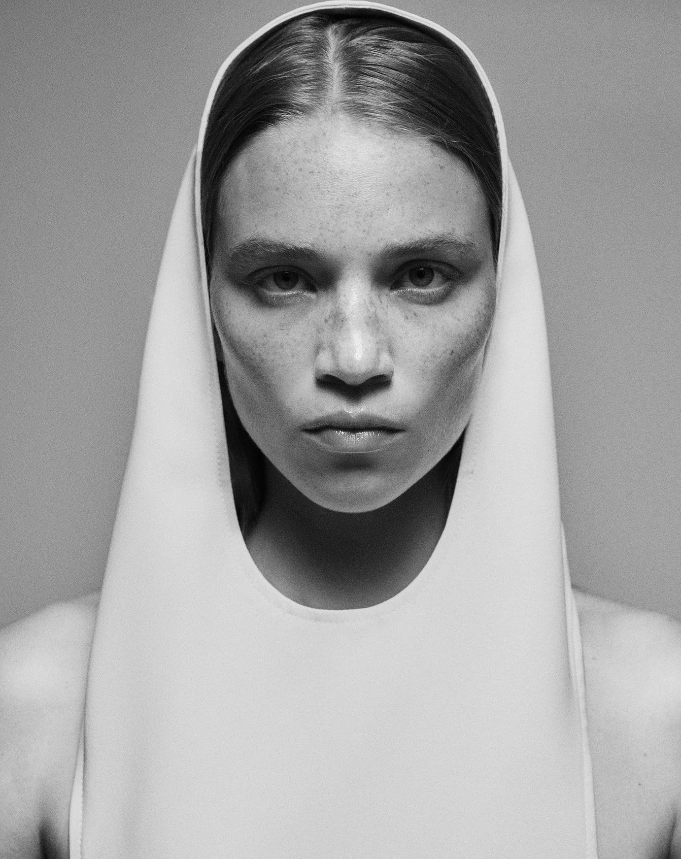 Black and white portrait of a young woman with straight hair, wearing a sleeveless top with a high neckline, and a white hood covering her head.