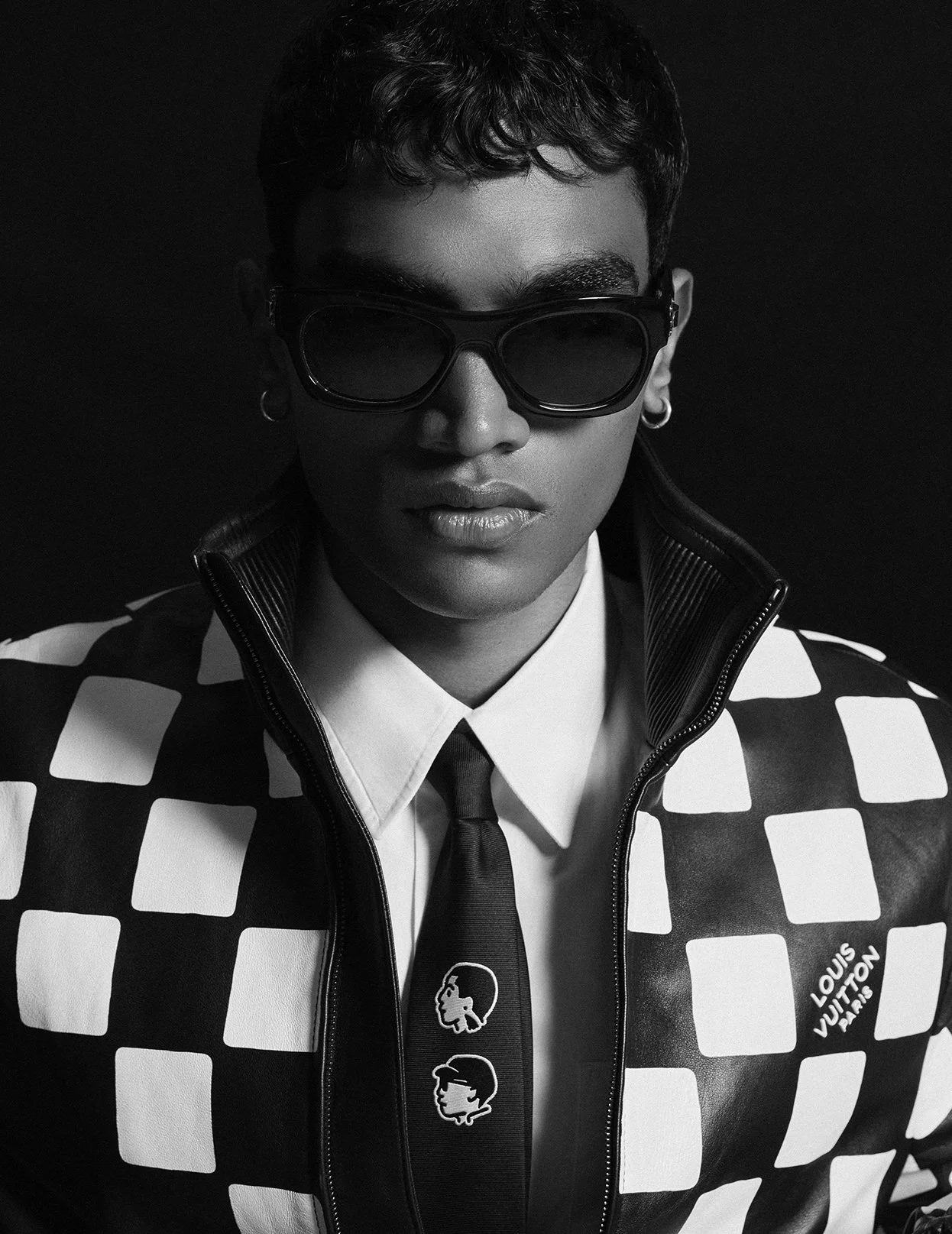 Black and white portrait of a person wearing sunglasses, a checkered Louis Vuitton jacket, white shirt, and a tie with cartoon face designs.
