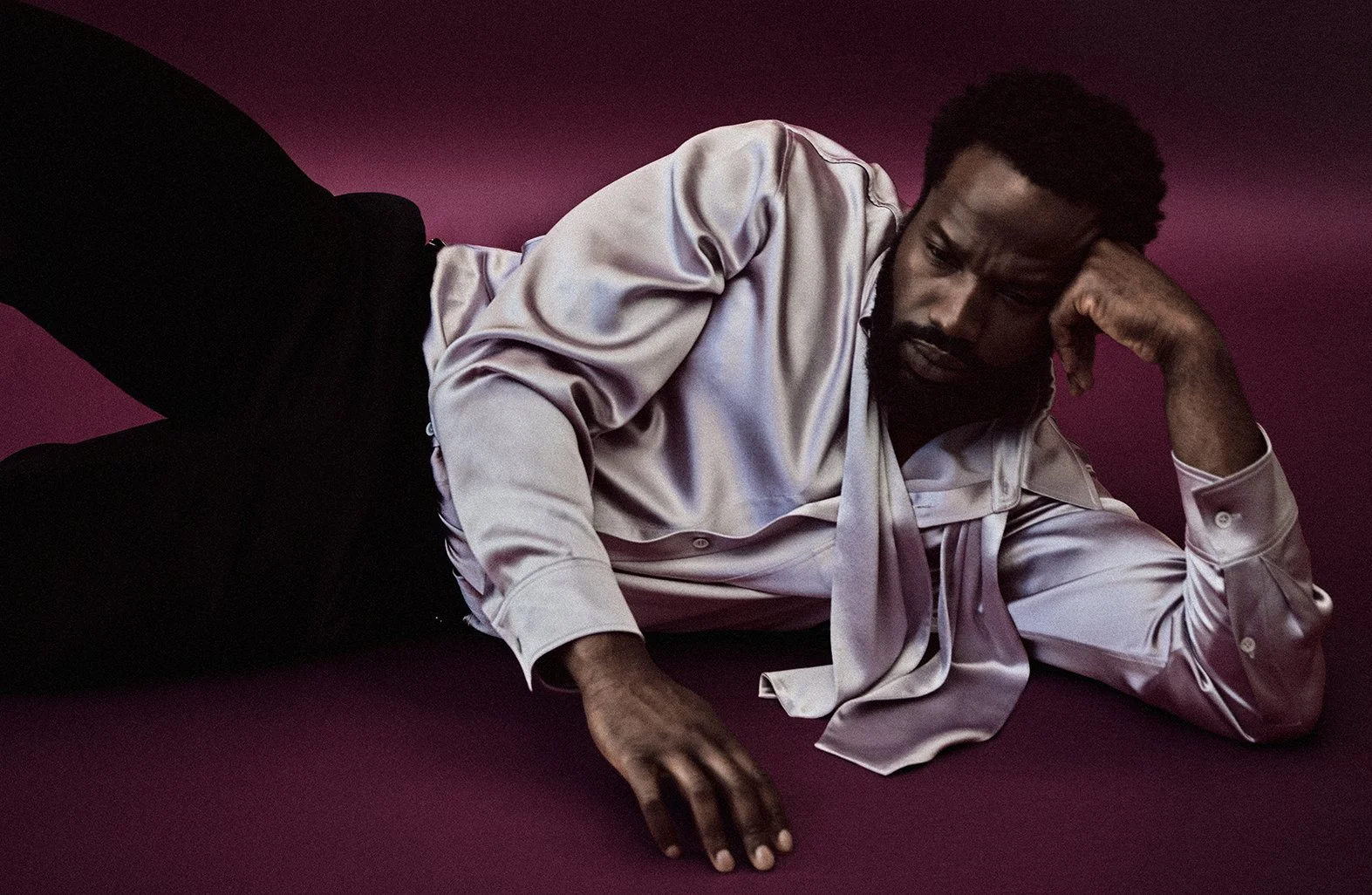 A man lying on his side on a maroon background, propped up on one elbow with a contemplative expression, wearing a shiny satin silver shirt and black pants.