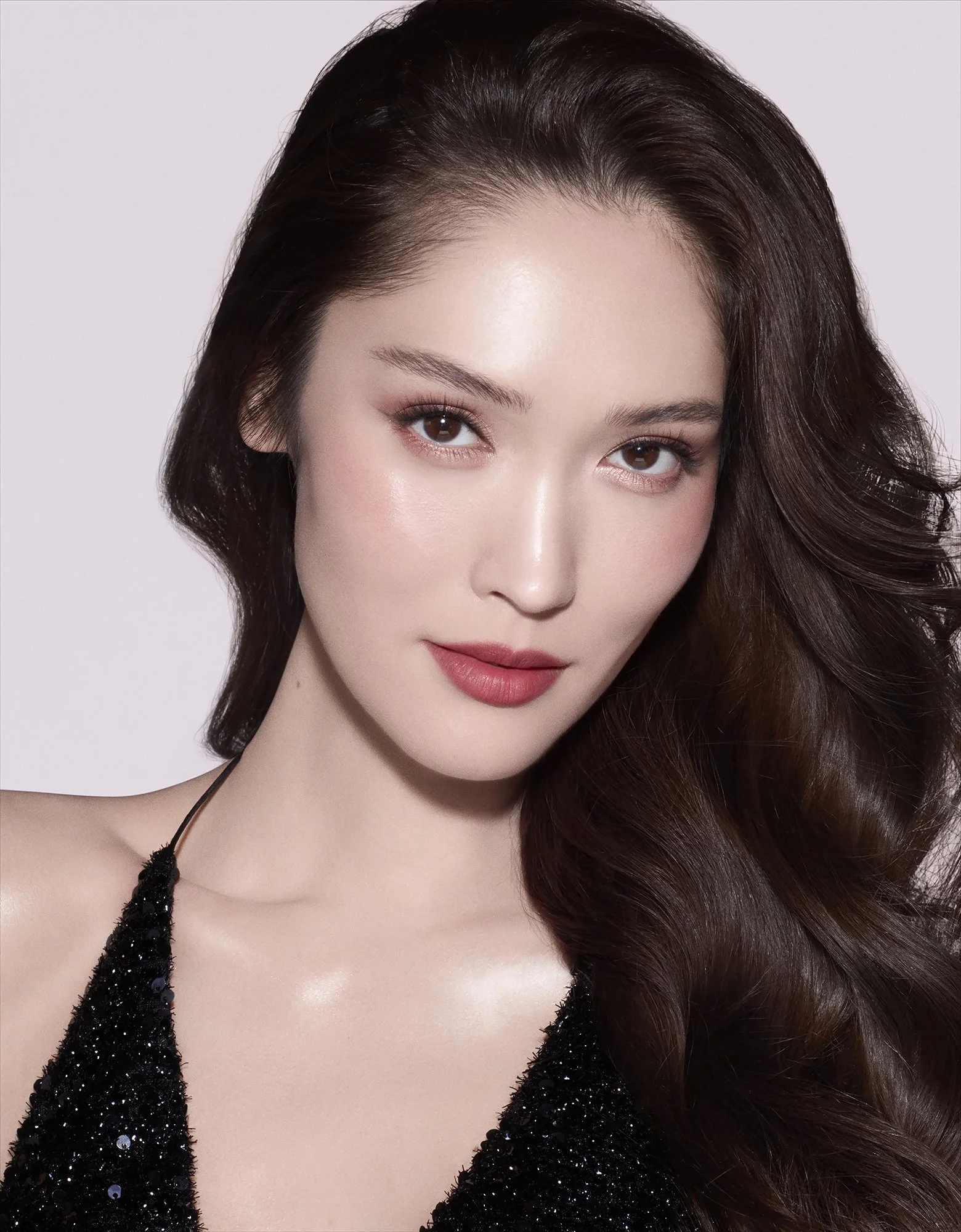 A close-up portrait of a young woman with long, wavy brown hair, fair skin, and subtle makeup, wearing a black, sparkly top against a plain light background.