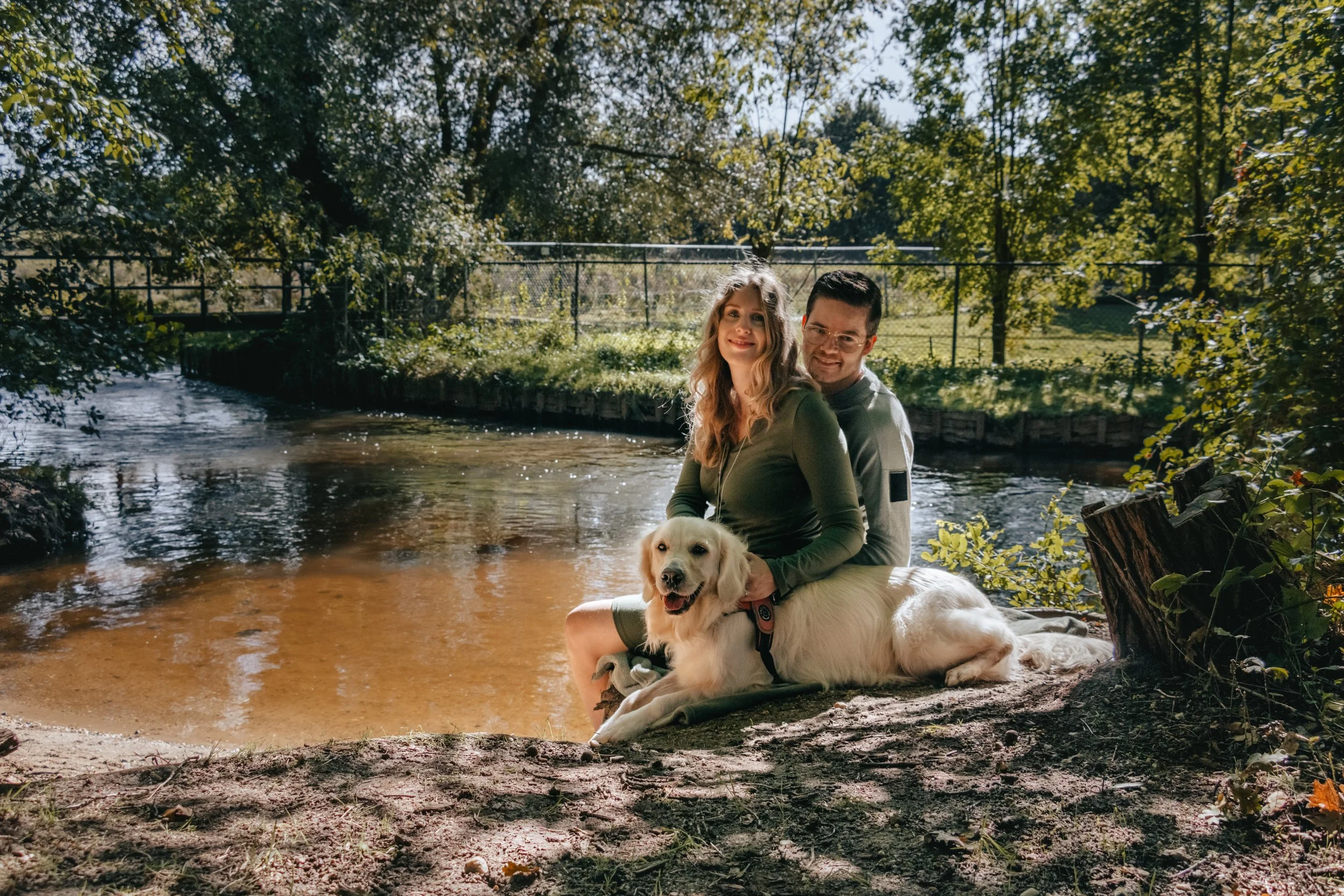 Family of three with father and pregnant mother with a dog sitting outside by the lake, surrounded by nature.