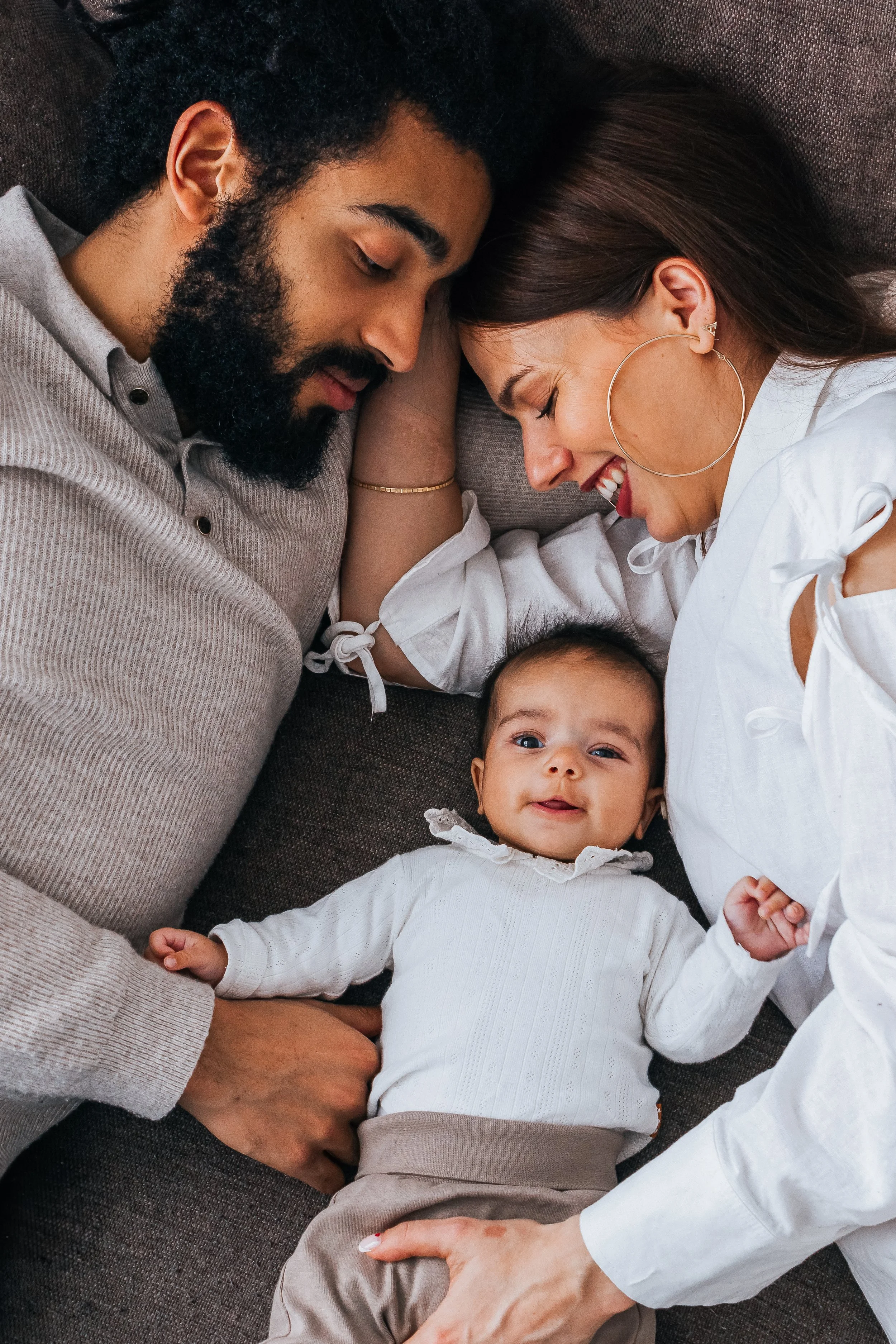 Family of three with a father on the left, baby in the middle and mother on the right lying down on the couch smiling towards their baby and baby looking to the camera.