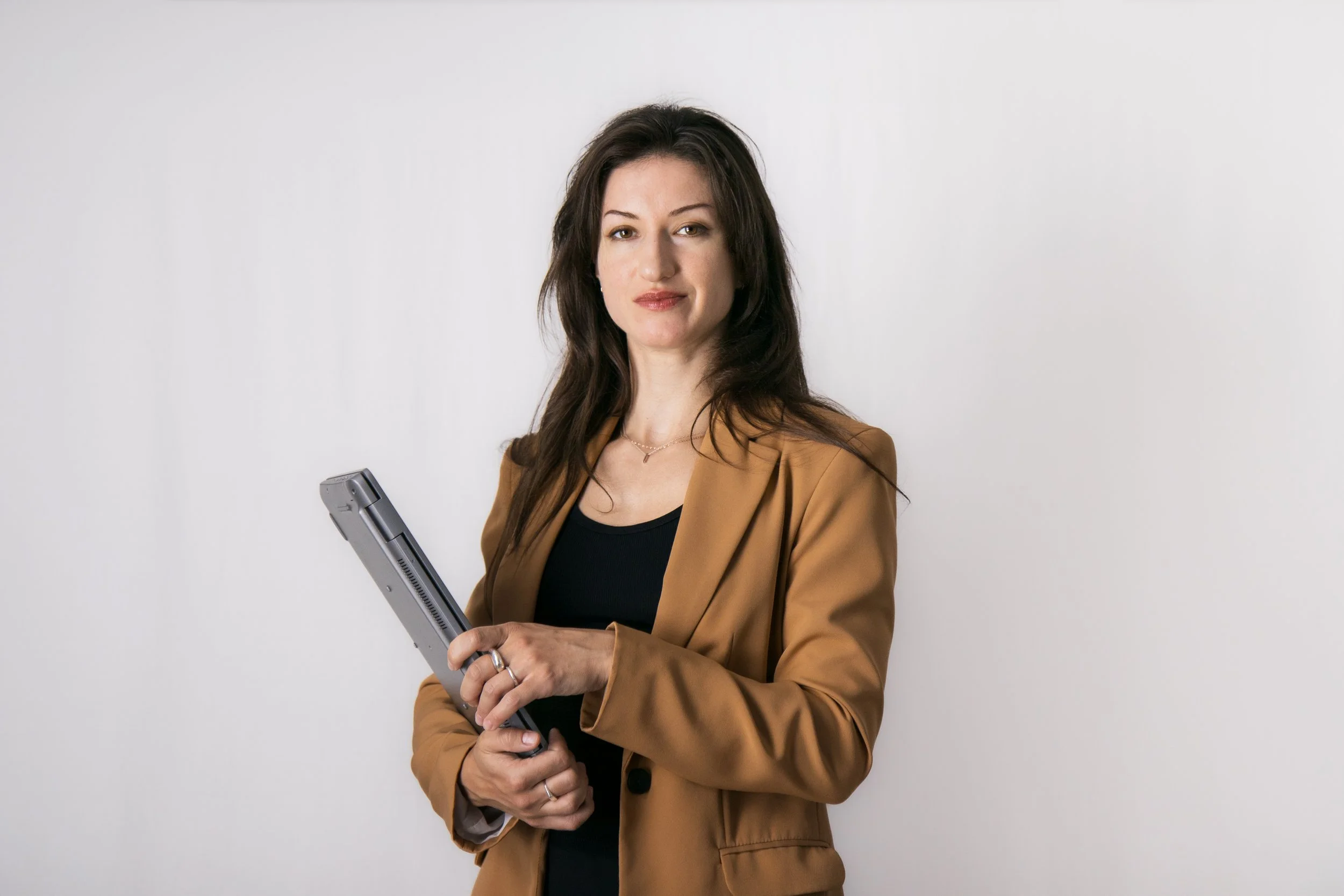 Woman standing in front of a white background with a blazer and a laptop, posing for a buisness profile picture