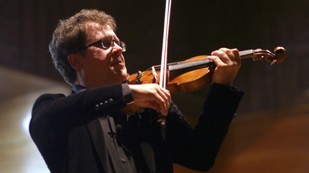 A man wearing glasses and a black suit playing a violin in a dimly lit performance space.
